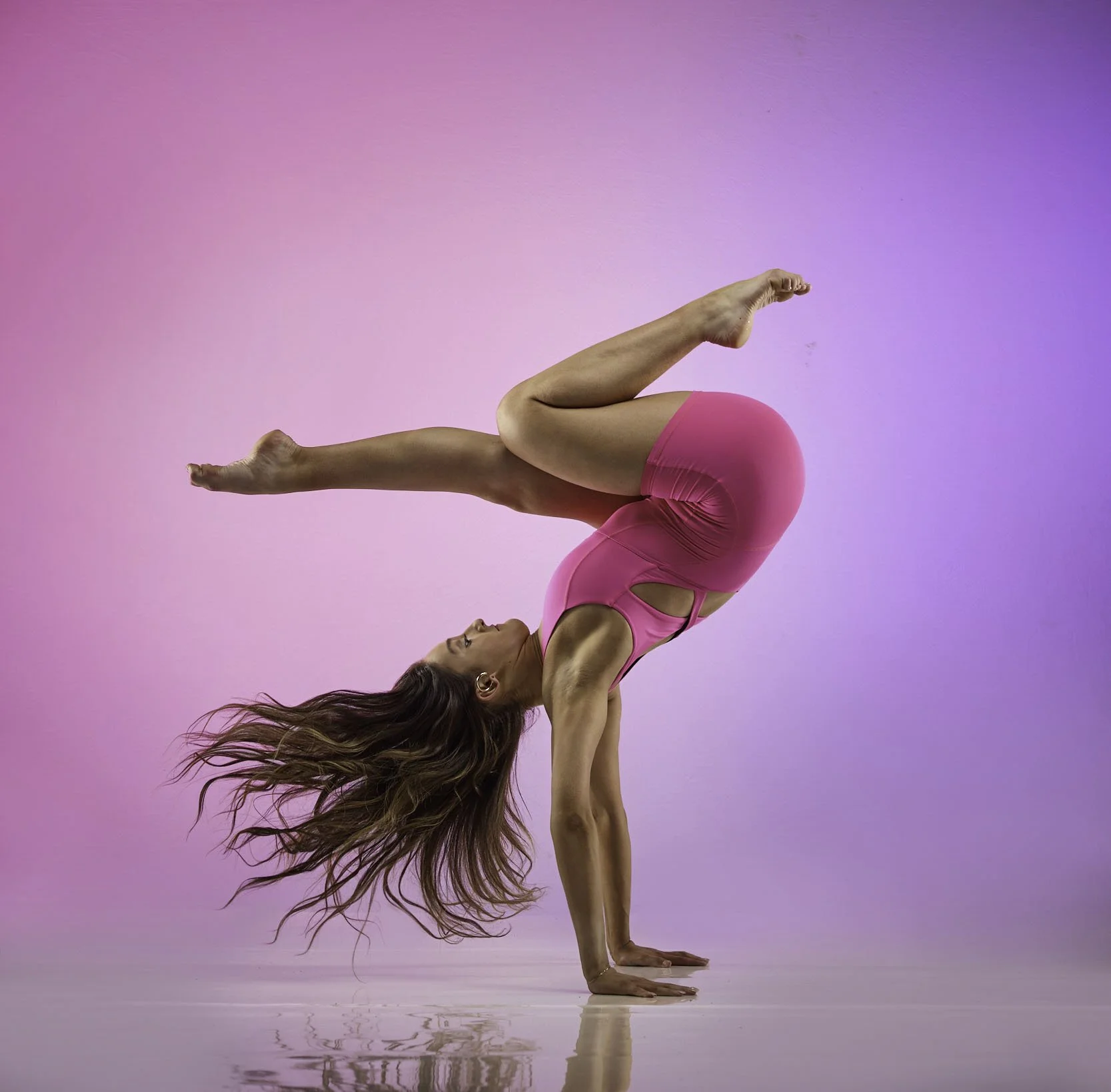 A woman in a pink athletic outfit performing a yoga pose called the headstand, with her legs bent and one foot resting on her opposite thigh, against a purple gradient background.