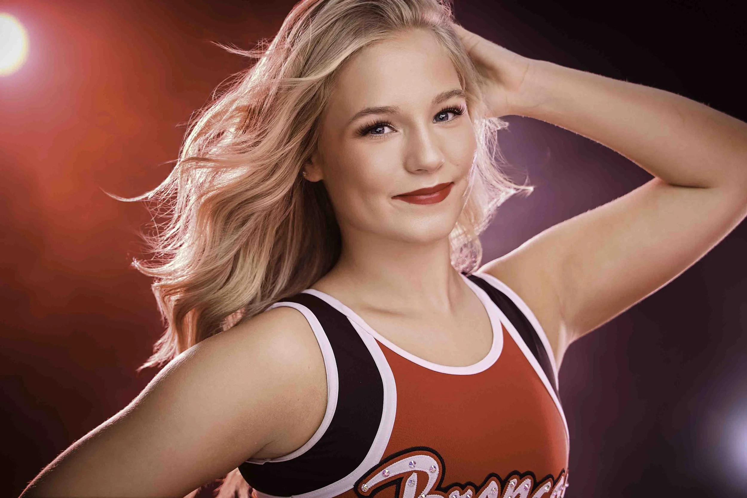 A young woman with blonde hair, wearing a red cheerleading outfit with black and white details, smiling and holding her arm behind her head, with a bright pink and purple background.