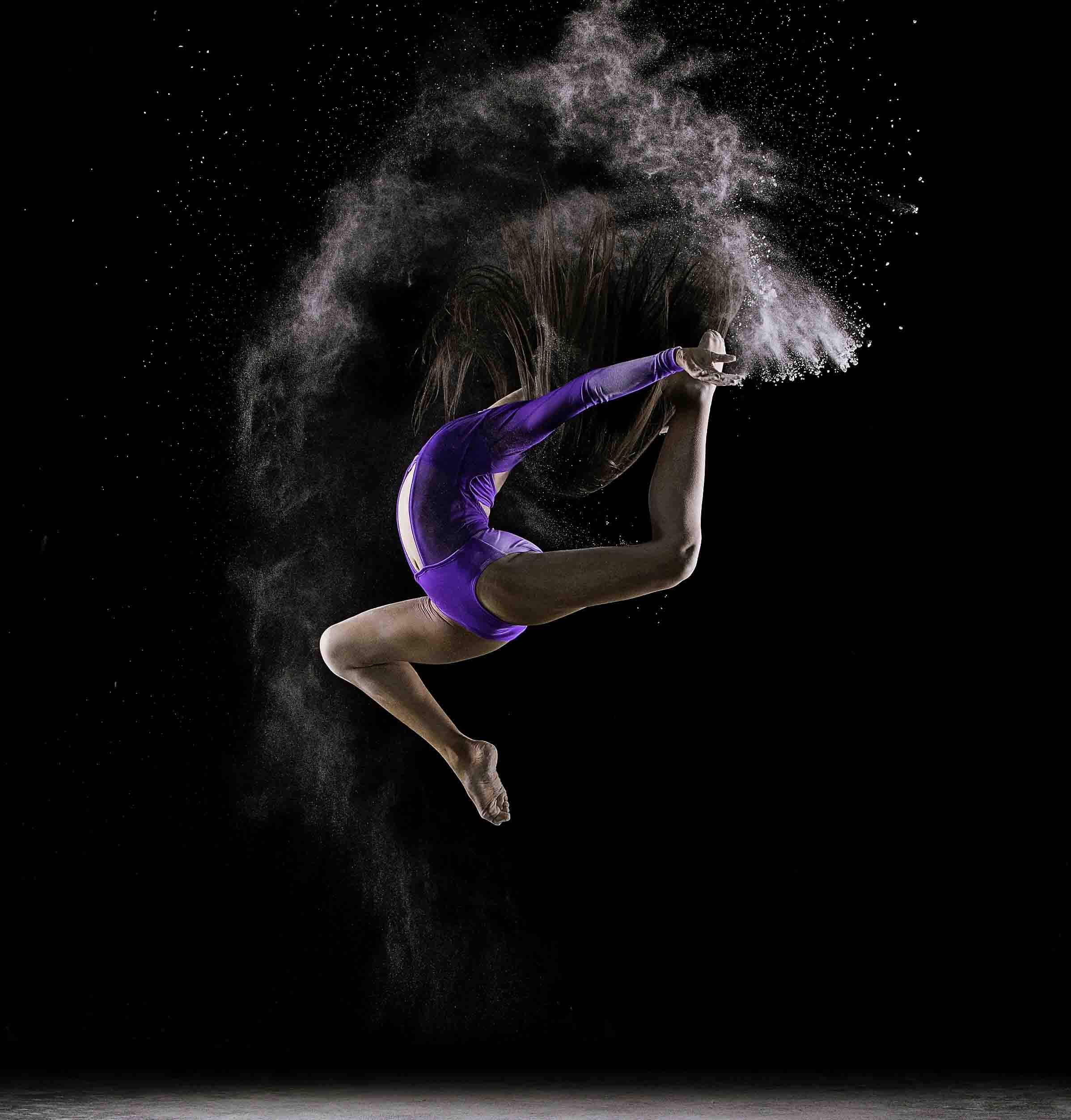 A woman in a purple sports outfit doing a backbend pose with pink-colored powder flying around her, set against a black background.