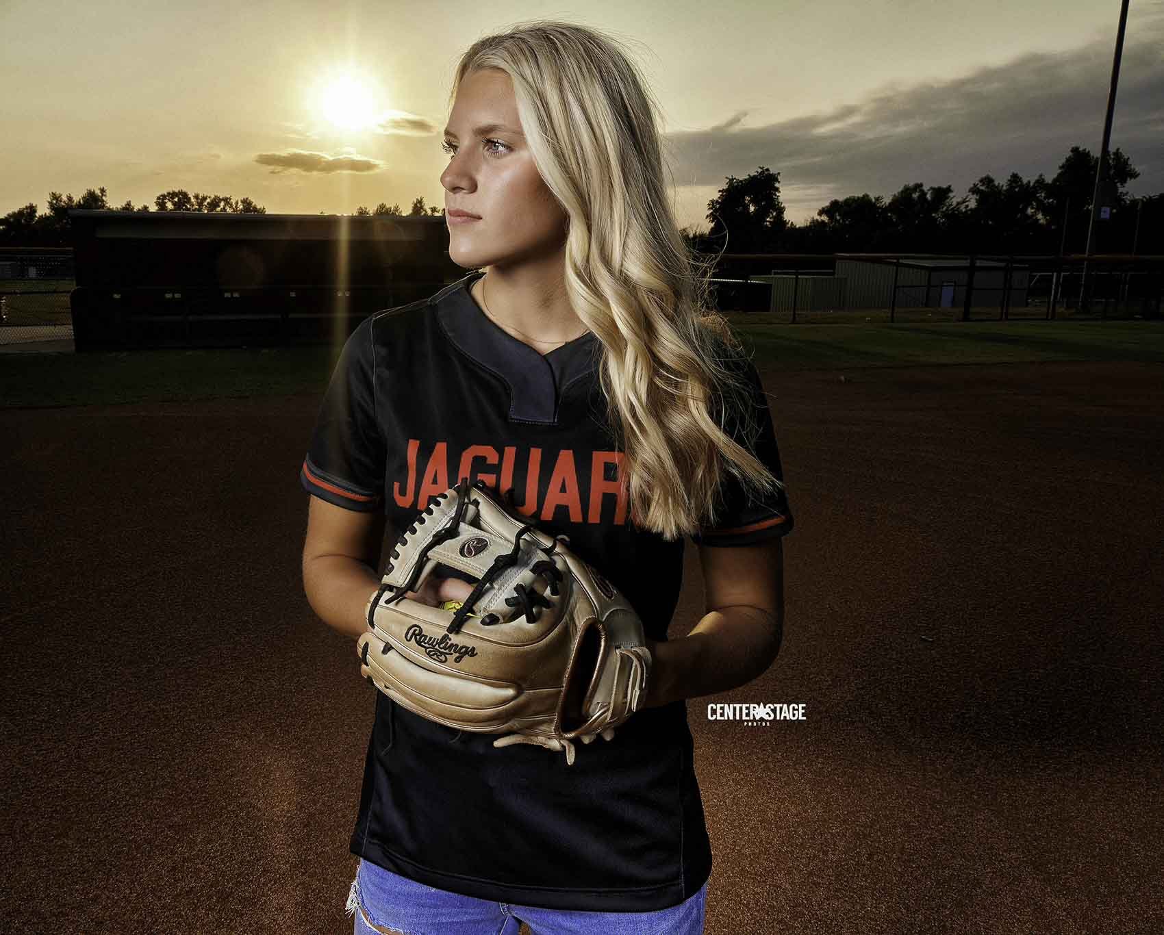 A young woman in a black sports jersey and blue shorts is standing on a baseball field during sunset, holding a baseball glove. She has long blonde hair and is looking to the side with a serious expression.