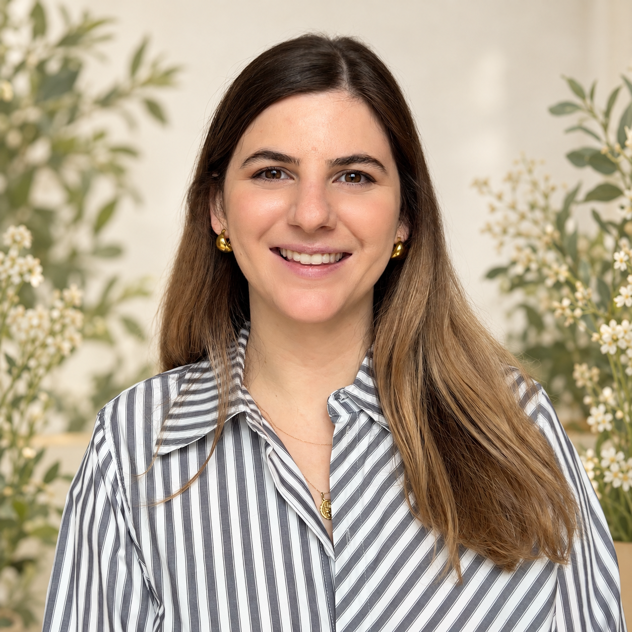 Brooke, a woman and licensed SLP with long brown hair wearing a striped shirt and gold jewelry, photographed indoors with flowering plants in the background