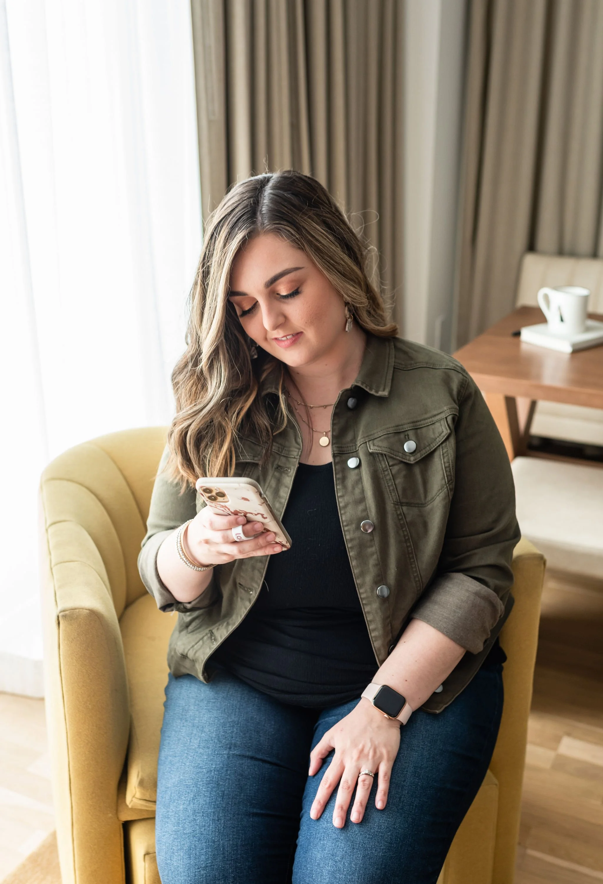 A woman with long wavy hair, wearing an olive green jacket, black top, and blue jeans, sitting on a yellow chair, looking at her smartphone.