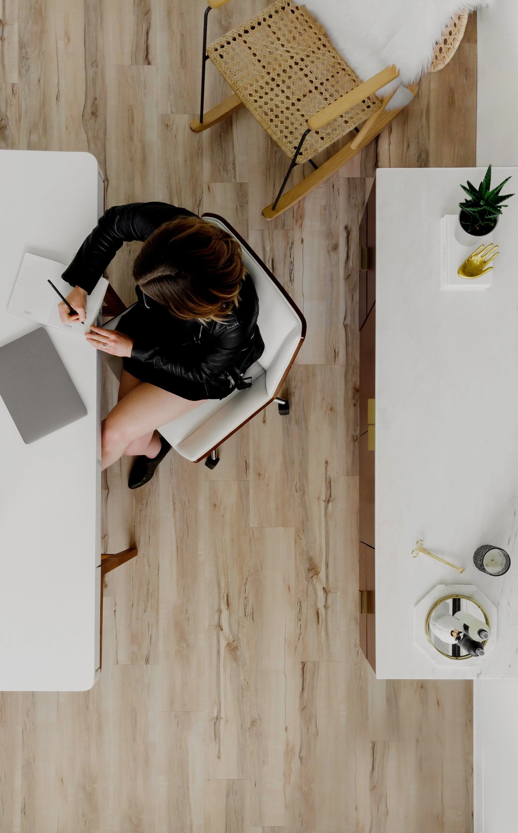 Top-down view of a woman sitting at a white desk, writing in a notebook with a laptop and pen nearby, in a modern room with wooden flooring and minimalist decor.
