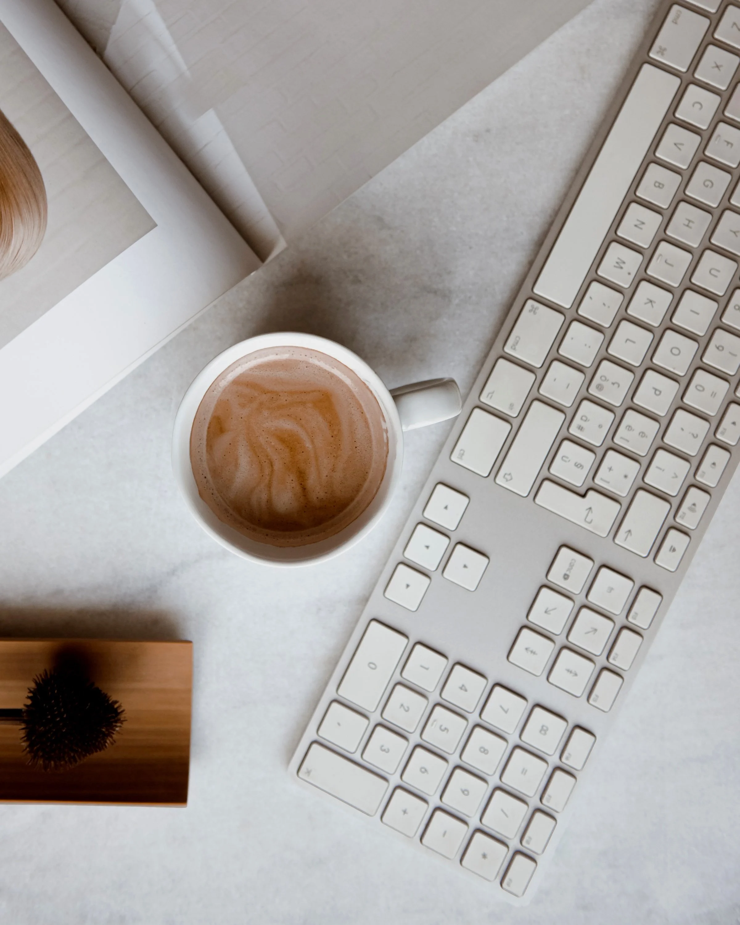 Computer keyboard, cup of coffee, book, and a small potted plant on a white desk.