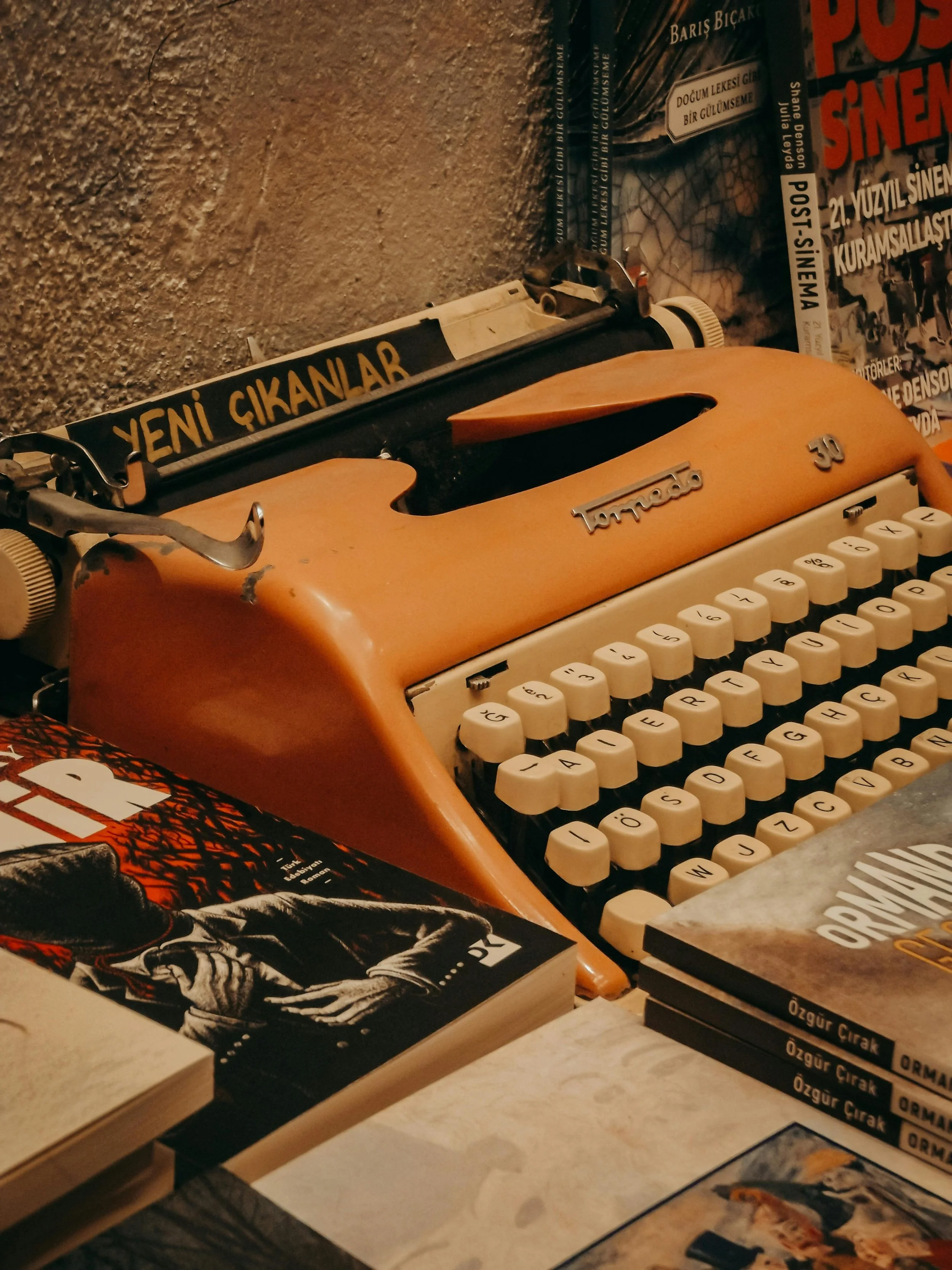 A vintage orange typewriter with white keys, surrounded by books and magazines on a cluttered table.