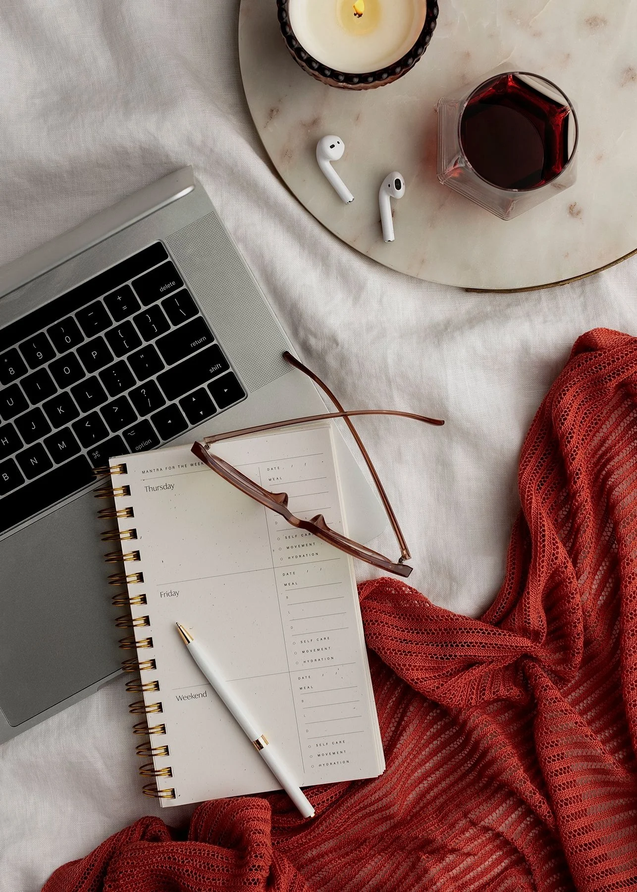 A flat lay of a workspace with a laptop, a weekly planner, a white pen, a pair of glasses, a marble tray with a candle, a glass of red wine, wireless earbuds, and a red blanket on a white surface.