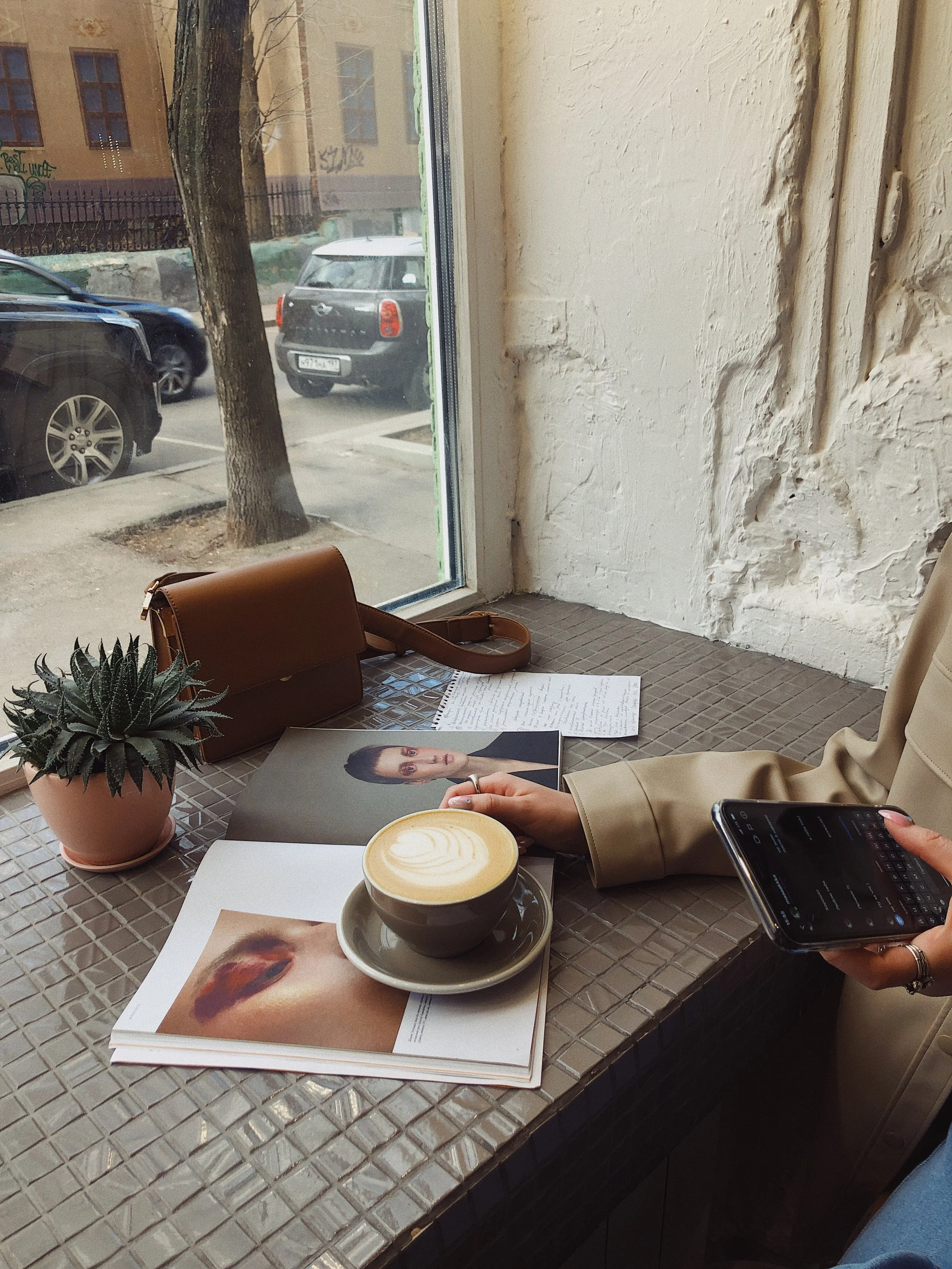 Interior of a cafe with a tray holding a latte, magazines, a purse, a plant, and a person using a smartphone, near a large window showing parked cars outside.
