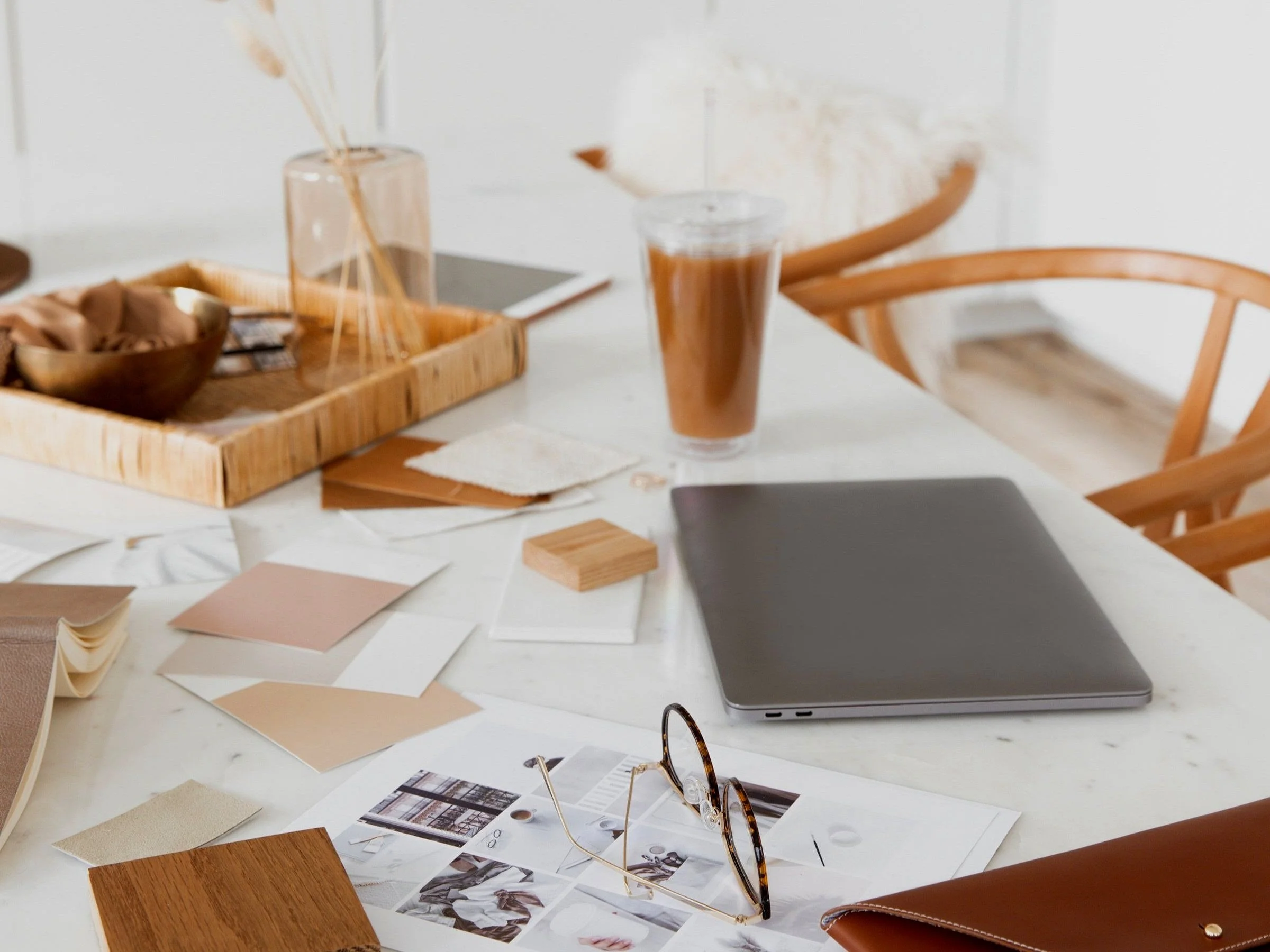 A white table with design and decor samples, eyeglasses, a laptop, a glass of iced coffee, and a brown paper bag, surrounded by wooden chairs in a bright room.