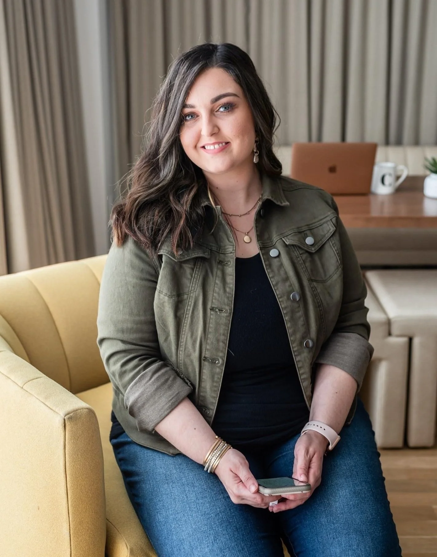 A woman with dark brown hair, wearing a green jacket and blue jeans, sitting on a yellow sofa and holding her phone in her hands. In the background, there is a wooden table with a laptop, a coffee mug, and a potted plant, with beige curtains behind.