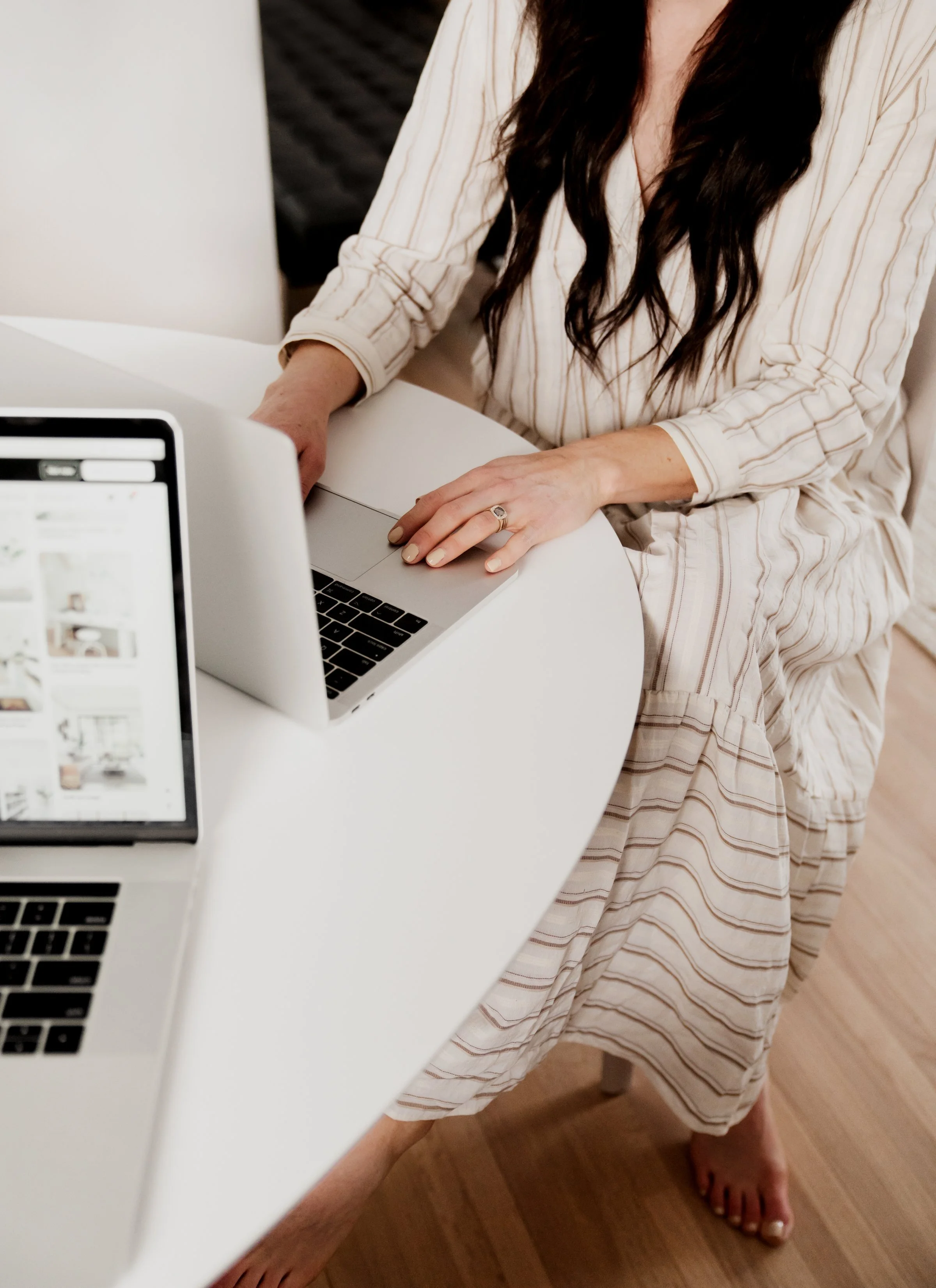 A woman with long dark hair working on a silver laptop at a white round table, with two other laptops visible, one showing a grid of images, in a cozy room with wooden floors and a bed in the background.