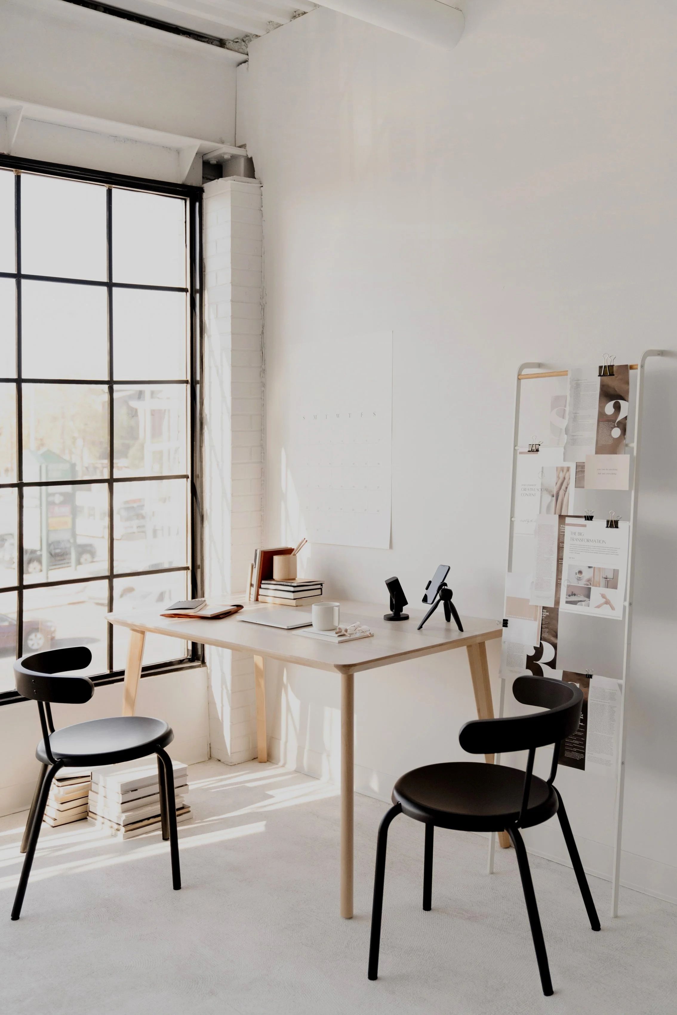 A bright, minimalist workspace with a large window, white walls, a wooden desk, two black chairs, a white bulletin board with papers, and a white calendar on the wall.