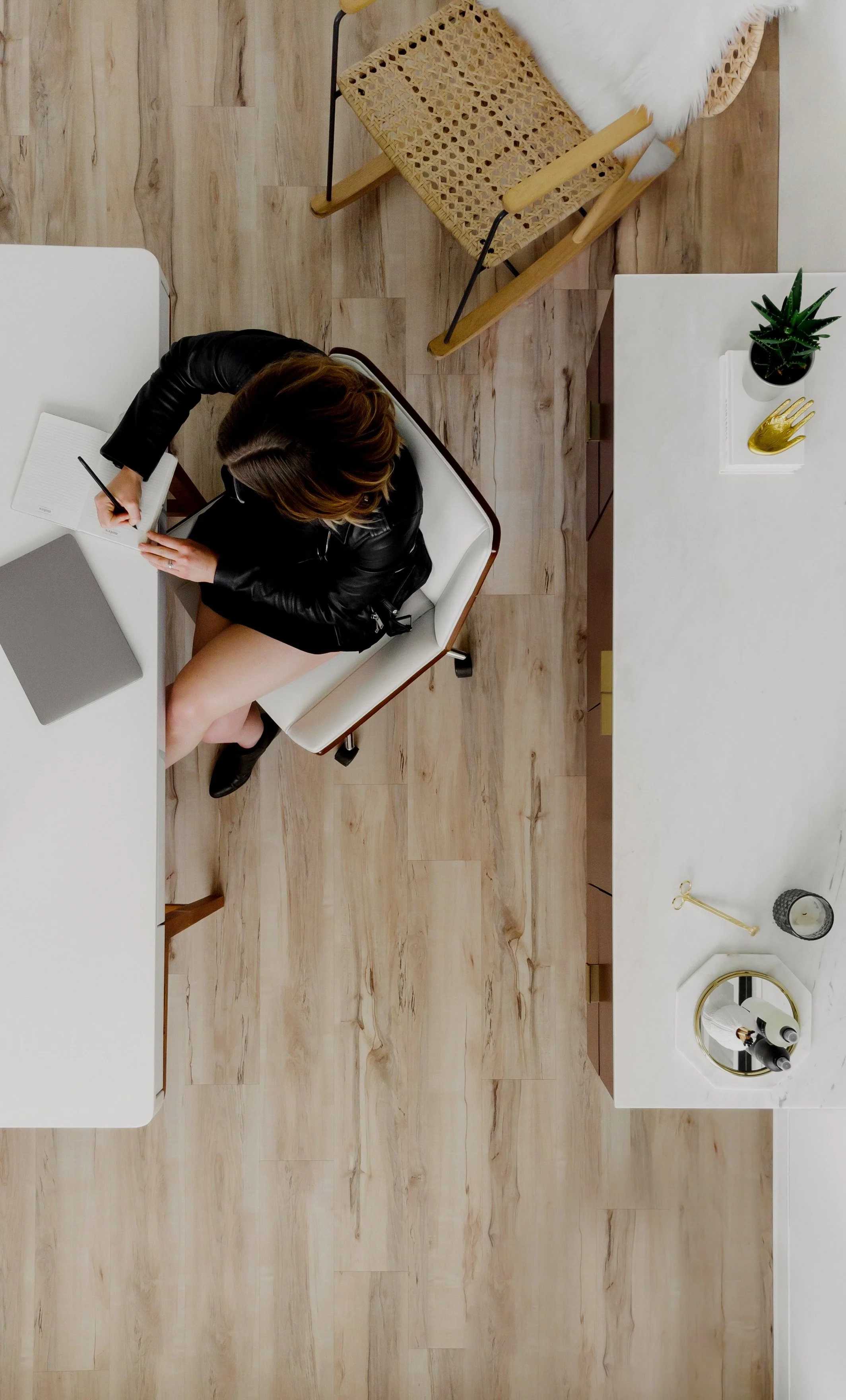 Top-down view of a woman sitting at a white desk, writing in a notebook, with a laptop nearby, in a modern room with wooden floor and a white marble top shelf with decorative items