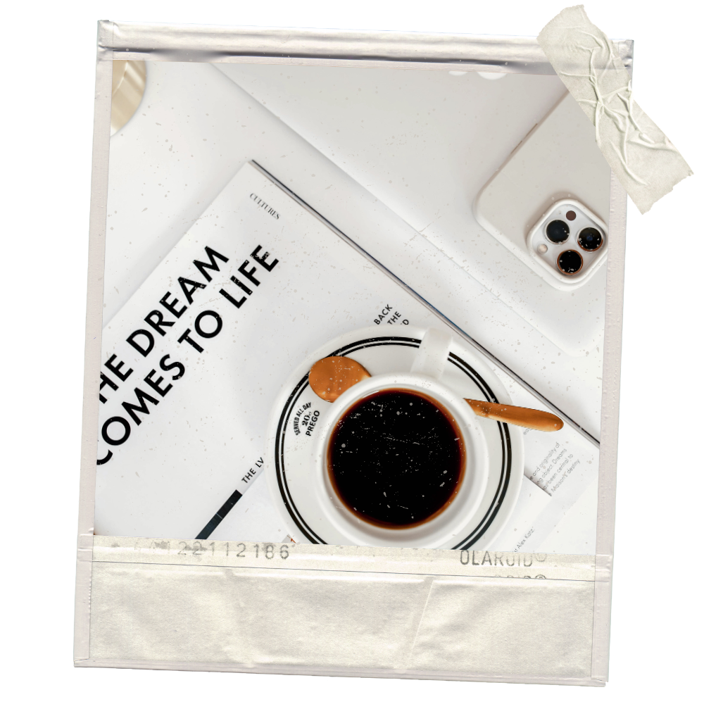 A cup of black coffee with a wooden spoon, a magazine titled "The Dream Comes to Life" on a white surface, a white smartphone with triple camera, and a crumpled tissue taped to the top right corner of the image.