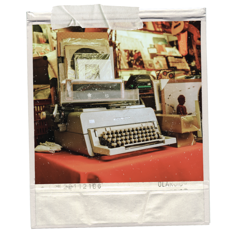 An old typewriter on a red table at a flea market or antique shop, with framed pictures and other vintage items in the background.