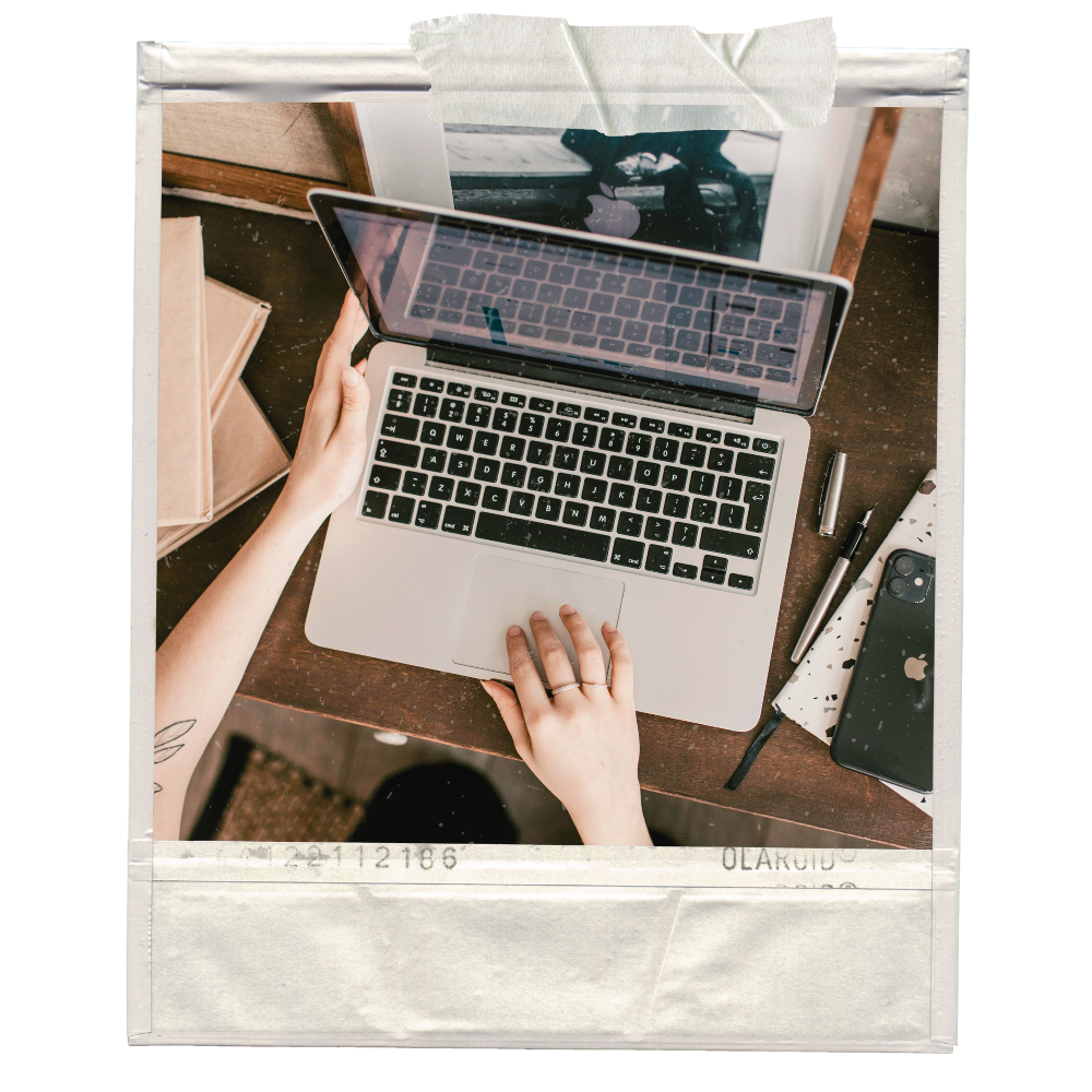 Top view of a person's hands working on a silver MacBook laptop on a wooden desk, surrounded by a pen, a notebook with a splattered paint design, an iPhone, and a marker, with a window in the background.