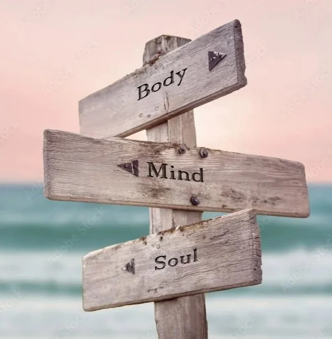 Wooden signpost with three arrows pointing in different directions, labeled "Body," "Mind," and "Soul," on a beach with the ocean in the background.