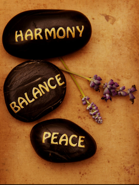 Three black stones with the words 'HARMONY', 'BALANCE', and 'PEACE' written in yellow, alongside a small bunch of purple lavender flowers, on a beige background.