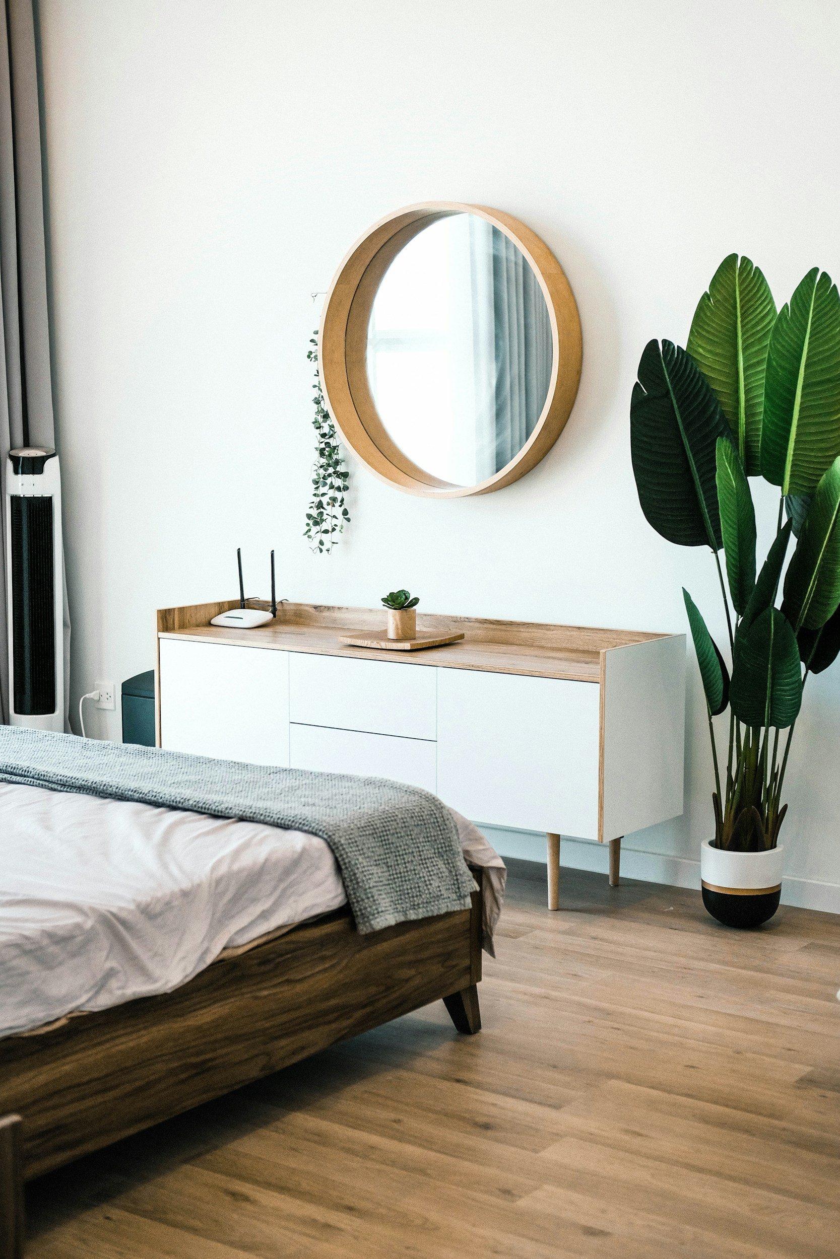 A bedroom interior with a bed, a wooden dresser with a mirror above it, a large potted plant, and a tall fan.