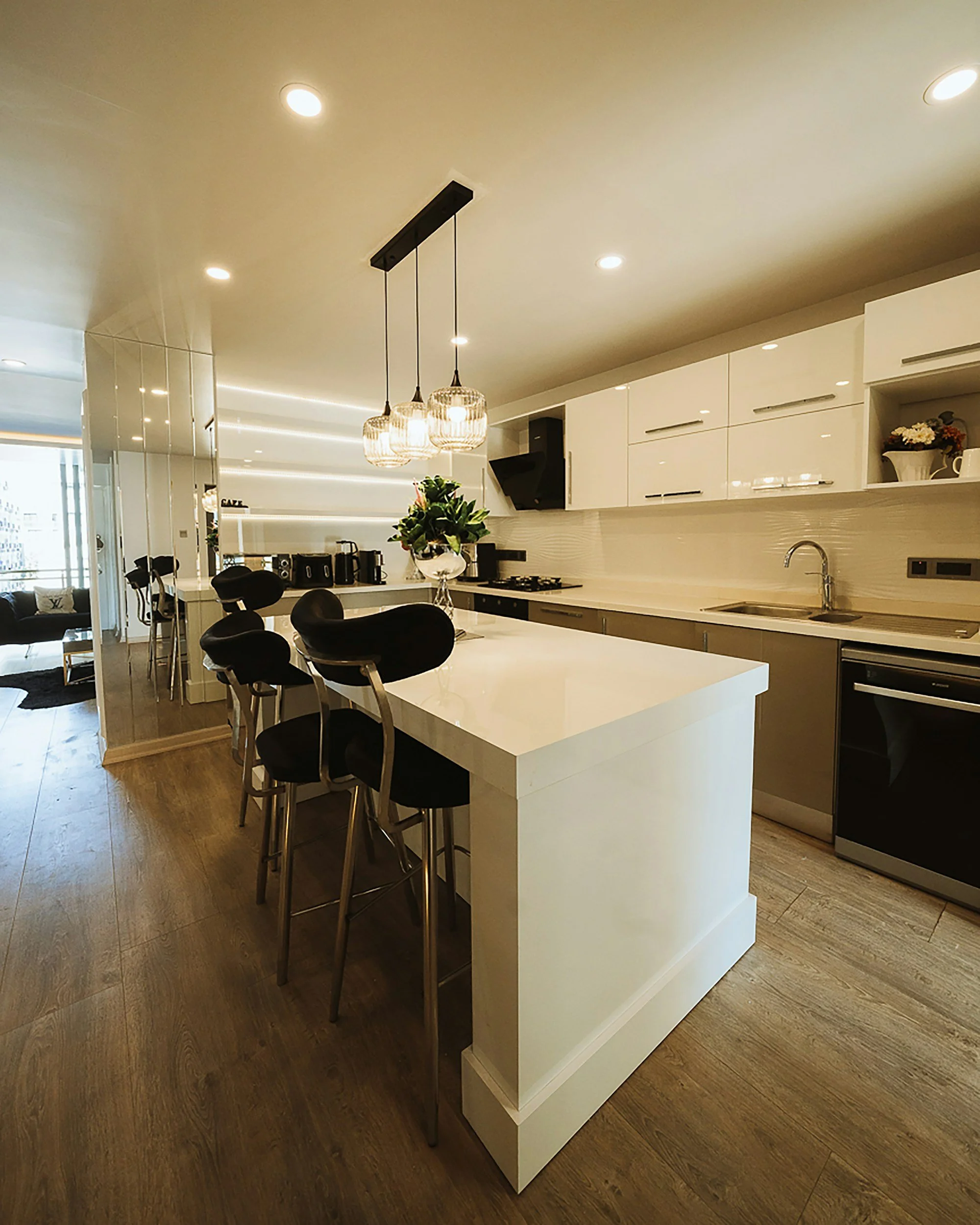 Modern kitchen with white cabinets, wooden floor, black chairs at a white kitchen island, pendant lights hanging from the ceiling, and a refrigerator.
