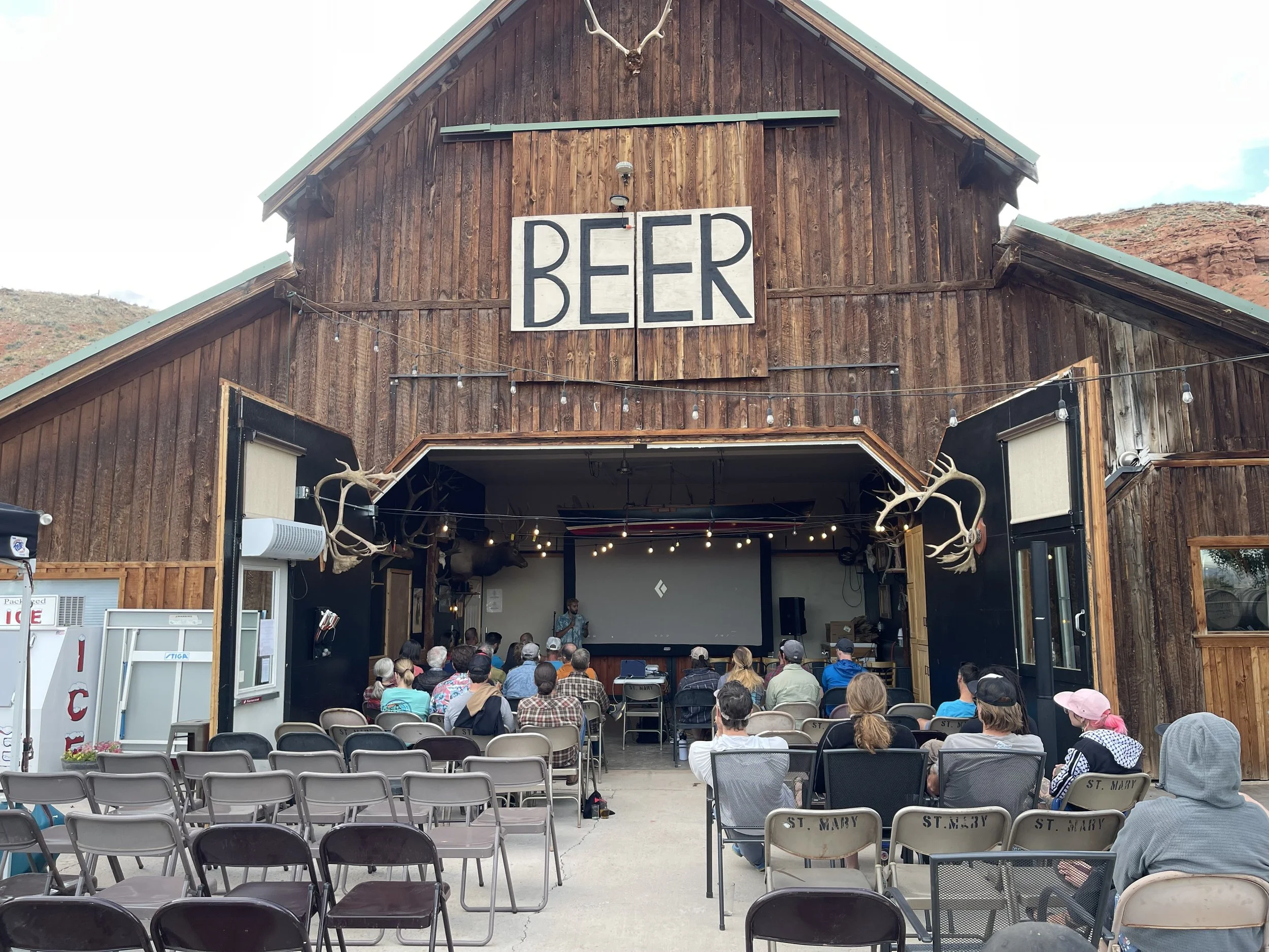 Audience watching a presentation in an outdoor venue with a rustic wooden building labeled "BEER" in the background. Some decorations include antlers mounted on the building's facade. The setting appears relaxed, with people seated on folding chairs, and a large screen at the front.