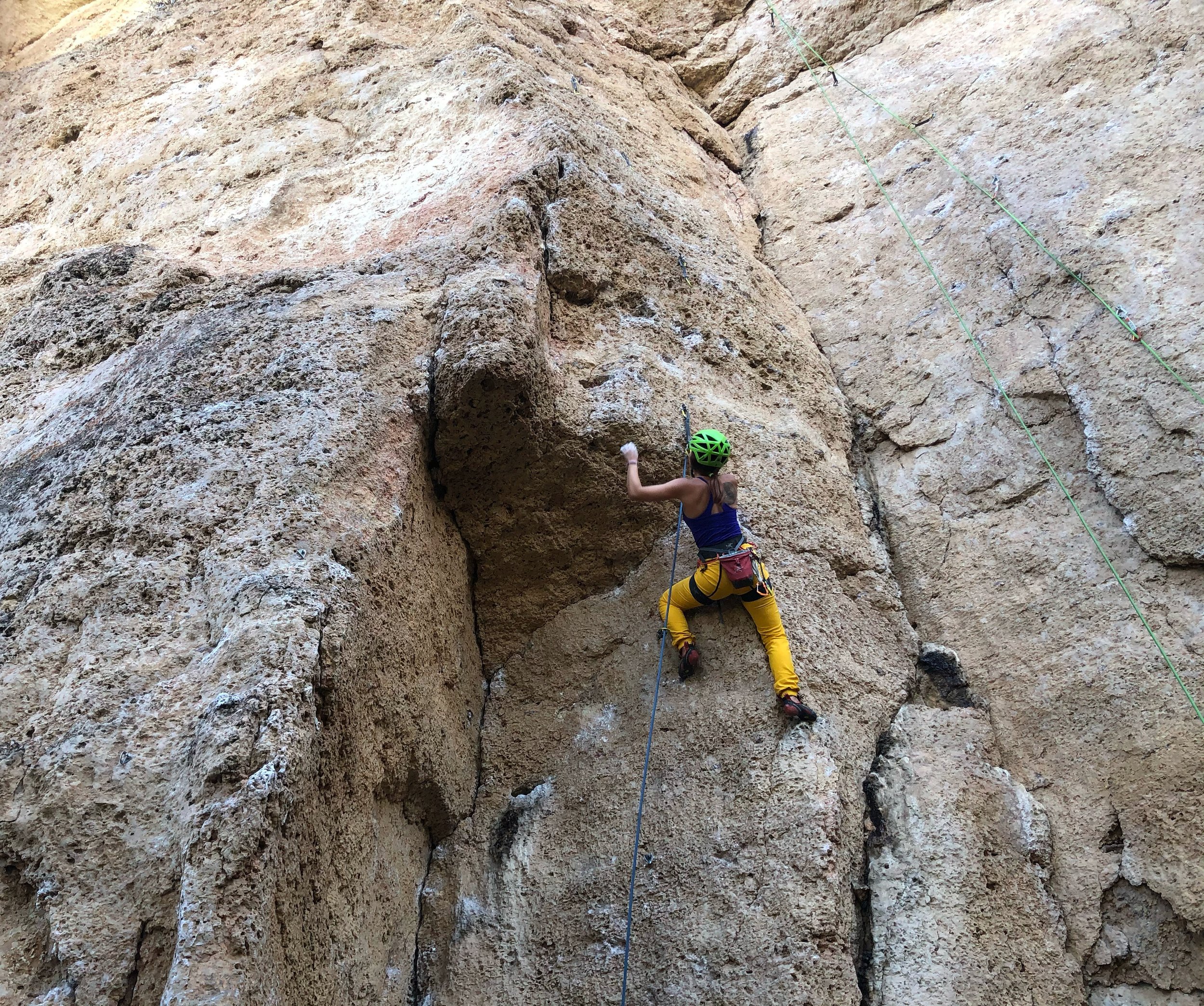 A woman rock climbing on a large, textured rock face, wearing a green helmet and yellow pants.