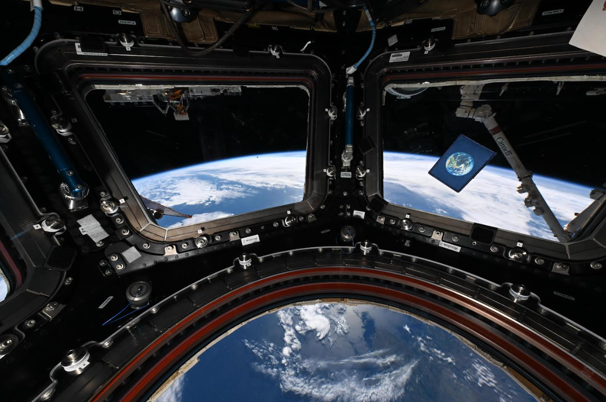 The Earth Flag on a notecard that is floating within the cupola of the ISS, with the Earth in the background as viewed through the windows