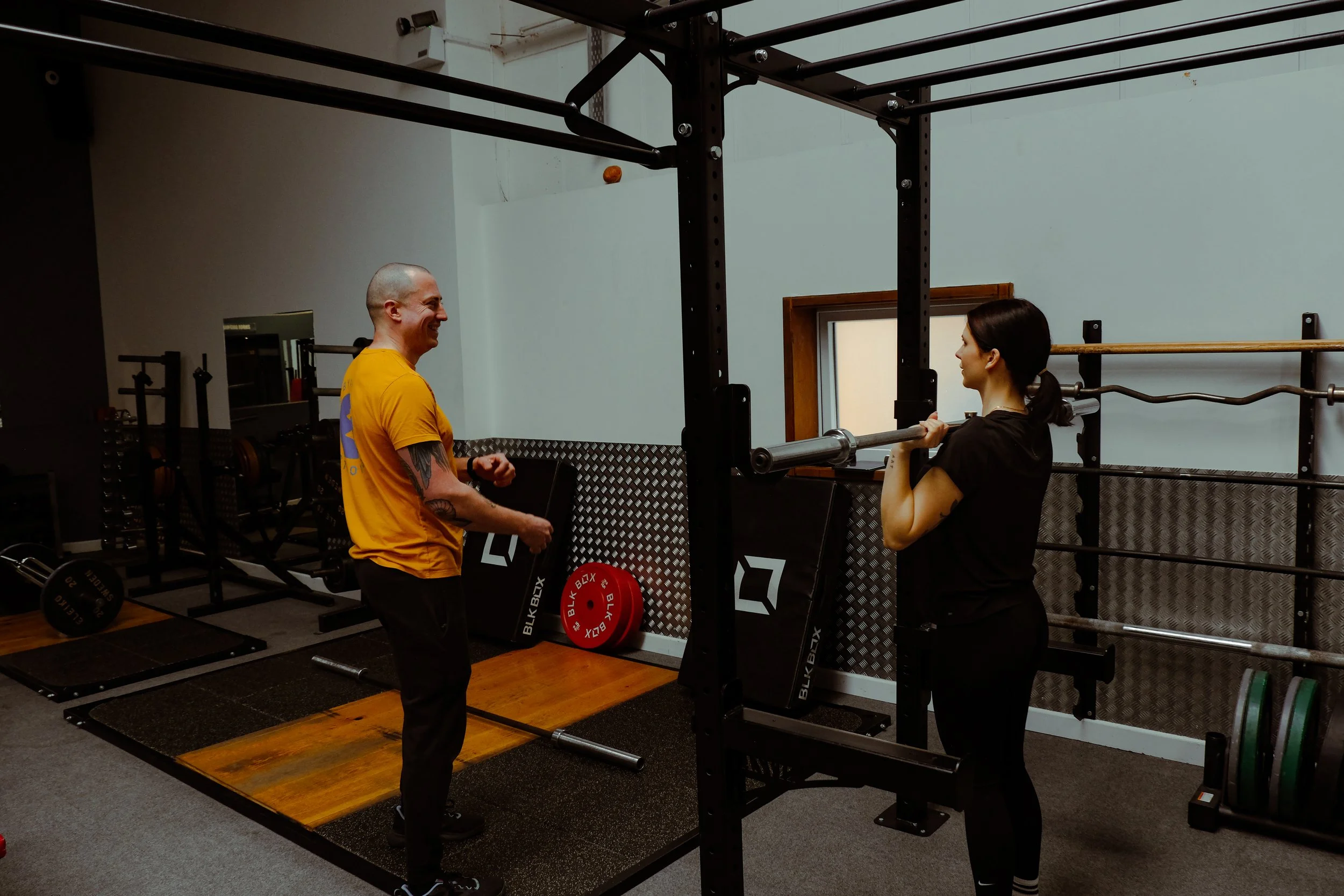 A man and a woman in a gym engaging in a fitness training session. The man is smiling and gesturing with his hands, wearing a yellow shirt. The woman is holding a barbell at shoulder height, preparing for or performing a lift, with a focused expression. The gym has weightlifting equipment, including barbells and weight plates.