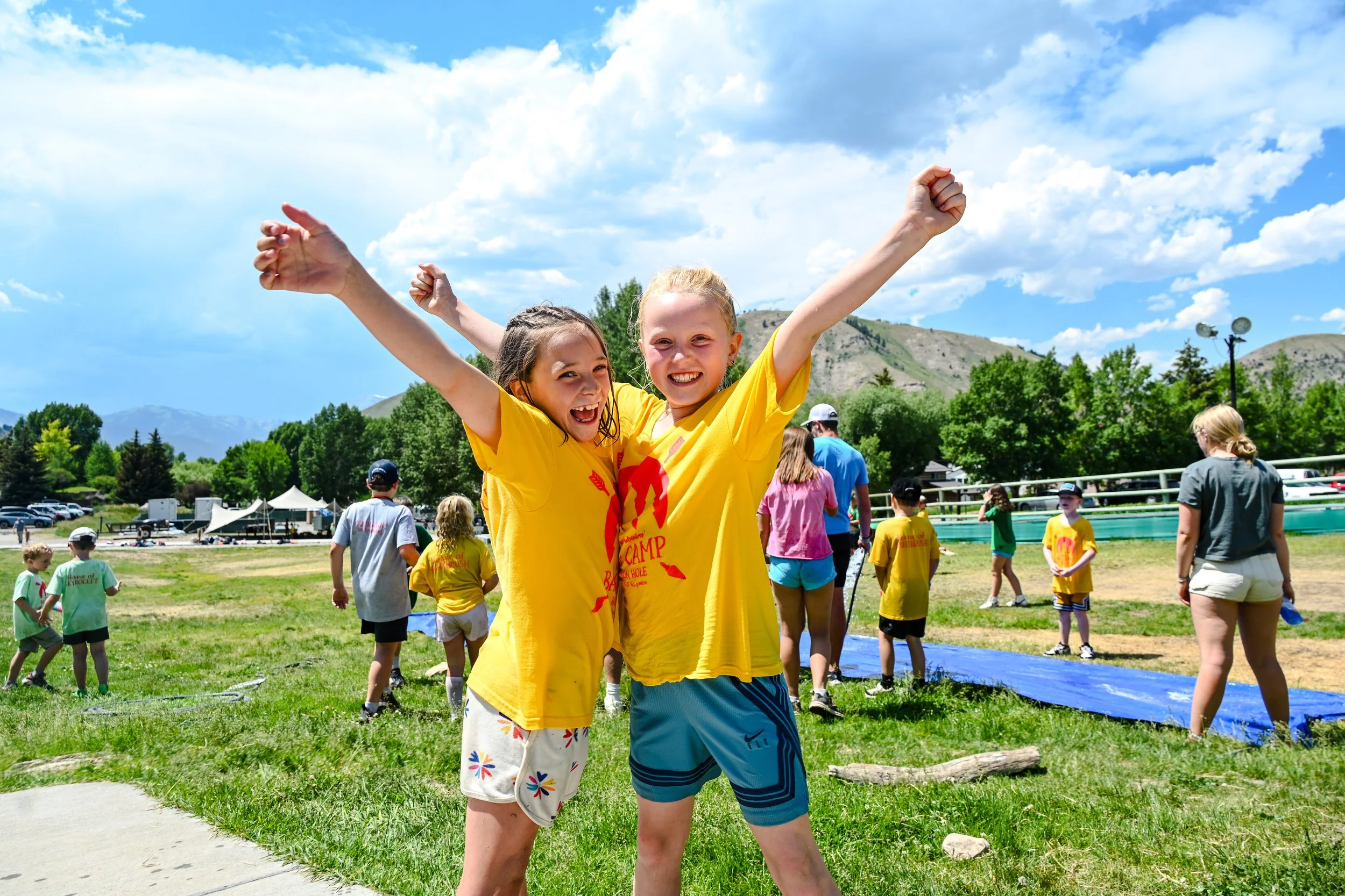 Two children celebrating outdoors at a summer camp with arms raised, smiling, and surrounded by other kids, trees, and mountains in the background.