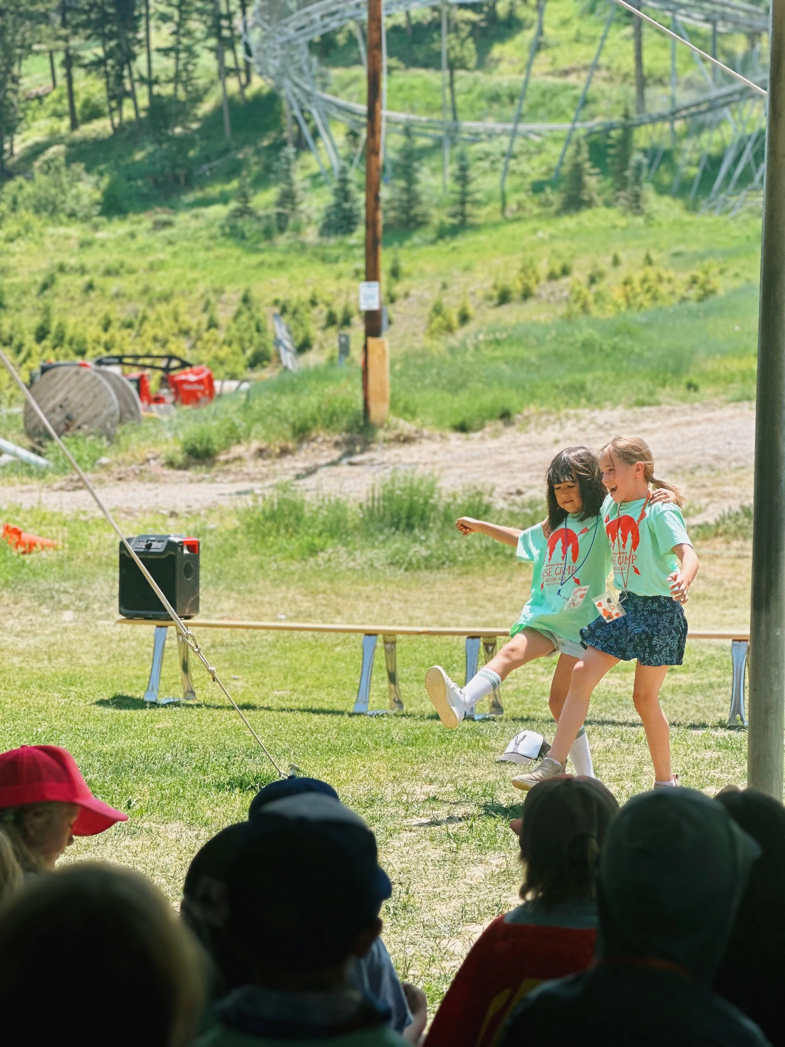 Two young girls dancing and playing on an outdoor stage during a summer camp, with children sitting in the foreground.