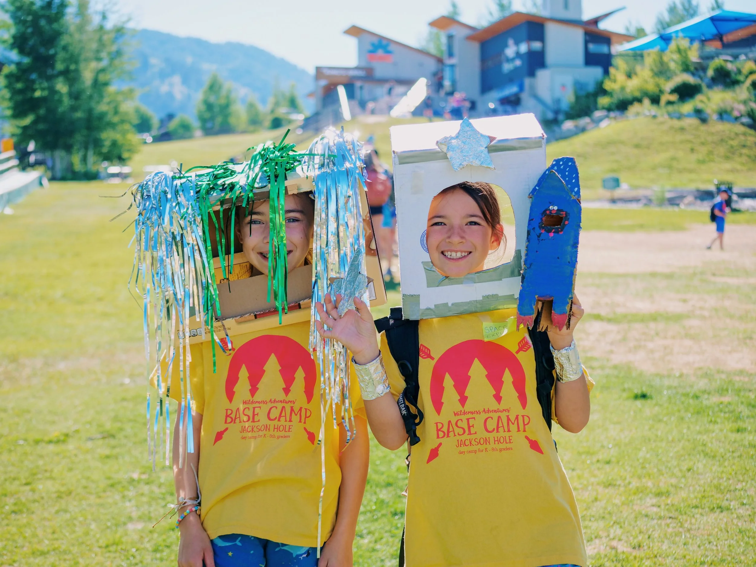 Two smiling children wearing yellow camp shirts with red trees and text, holding cardboard boxes decorated to resemble space helmets with metallic fringe and a star ornament, standing outdoors in a grassy area with trees and buildings in the background.