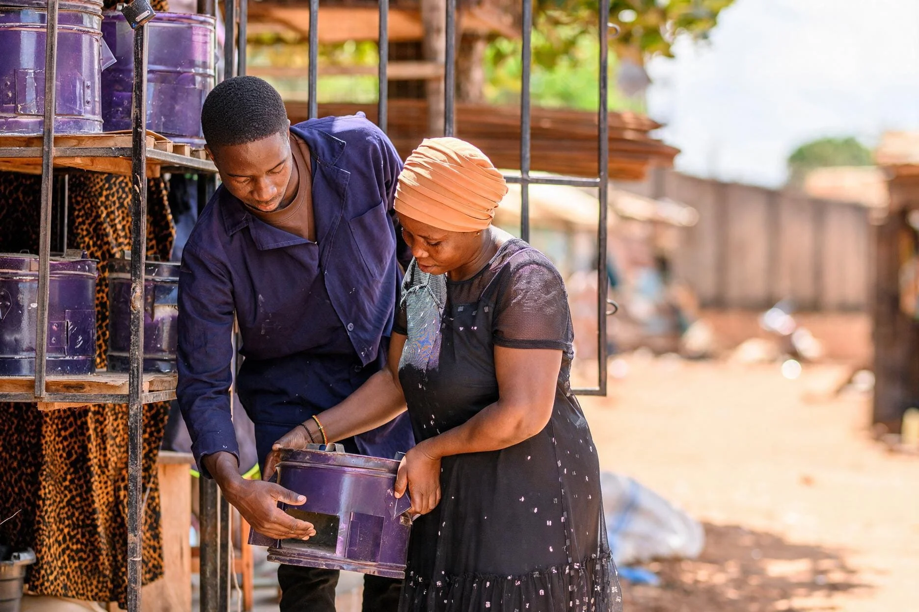 A man and woman looking at a purple container outdoors, surrounded by shelves of similar containers, in a marketplace setting.