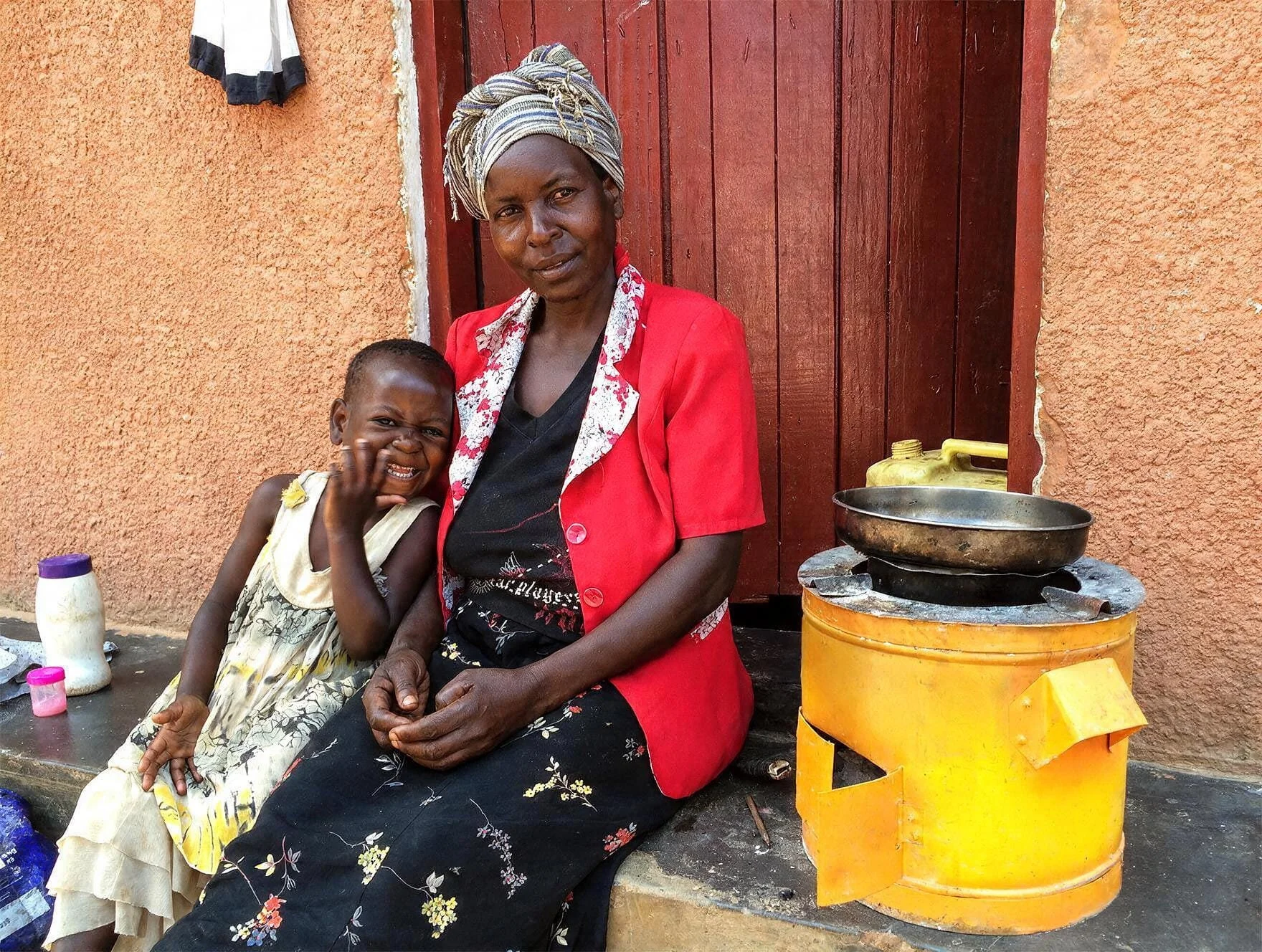 A woman and a young girl sitting outside next to a yellow stove, against a textured orange wall and a wooden door.