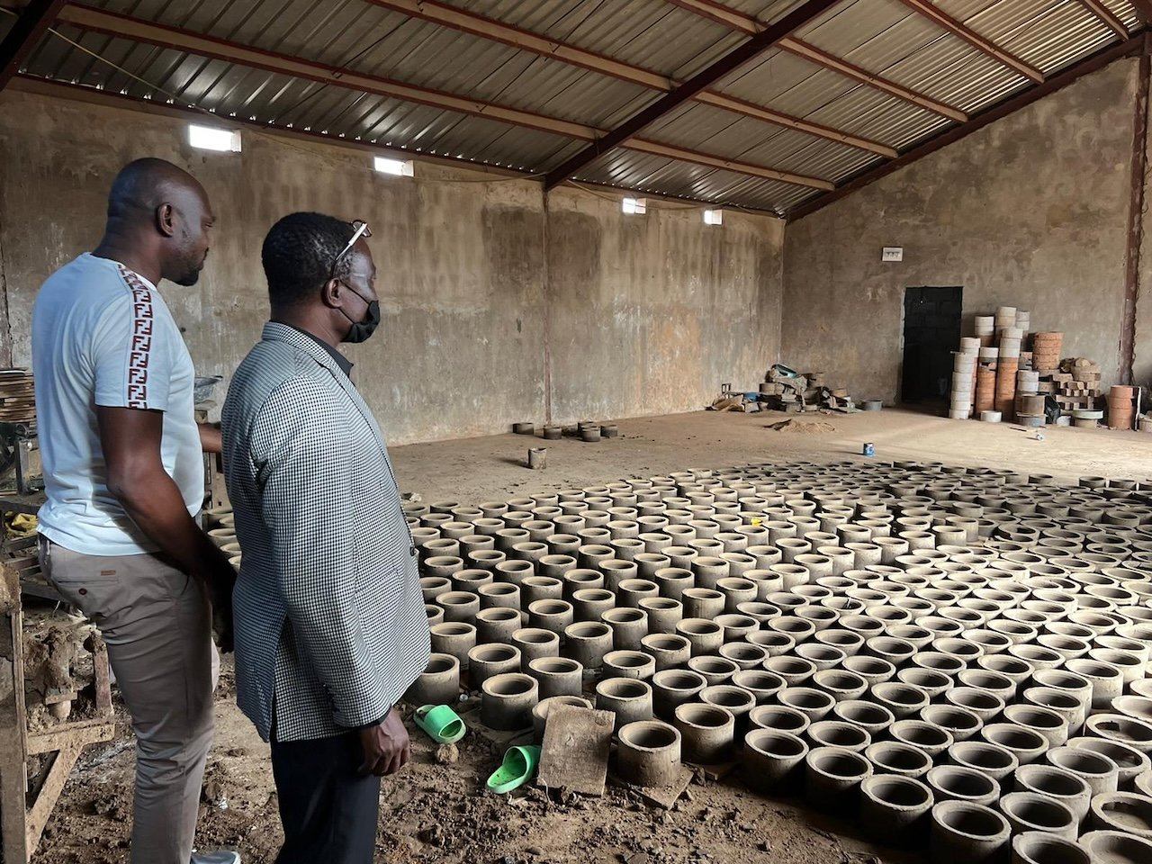 Two men examining a large number of small clay pots inside a warehouse or workshop. The workshop has a metal roof and unfinished walls, with stacks of additional clay pots in the background.