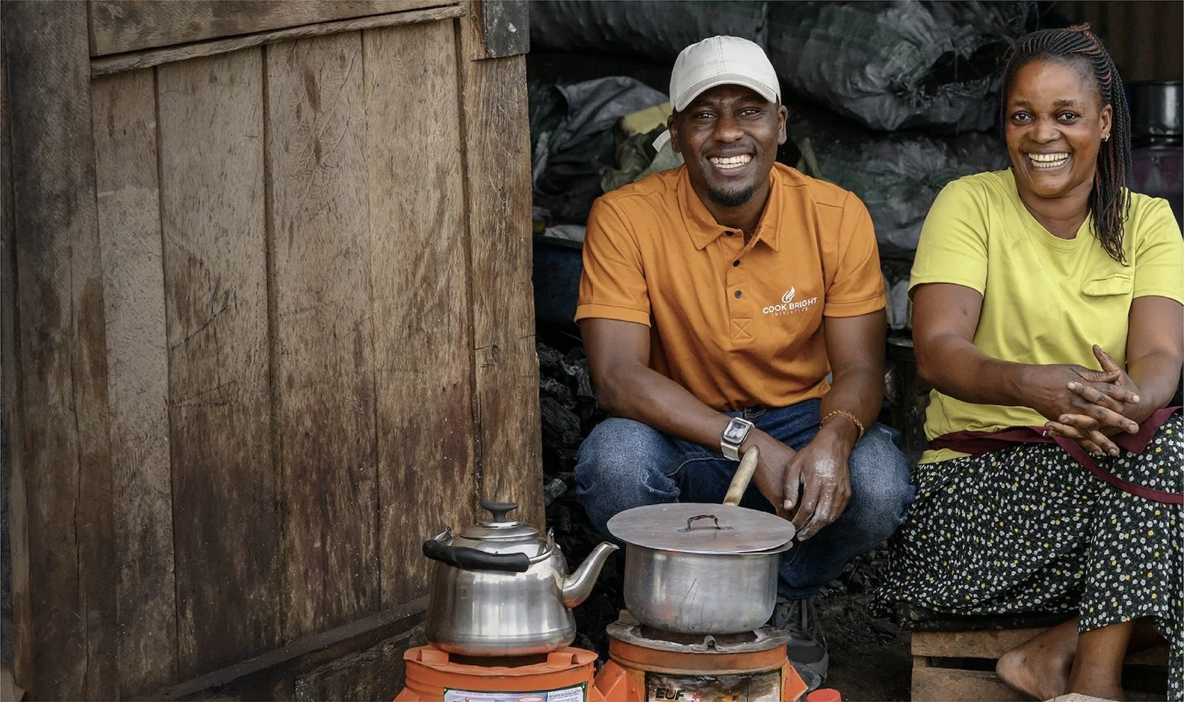 A man and woman sitting inside a simple wooden structure, smiling for the camera. The man is wearing a white cap and an orange shirt with a logo, and is sitting behind a improved cookstove with a stainless steel kettle.