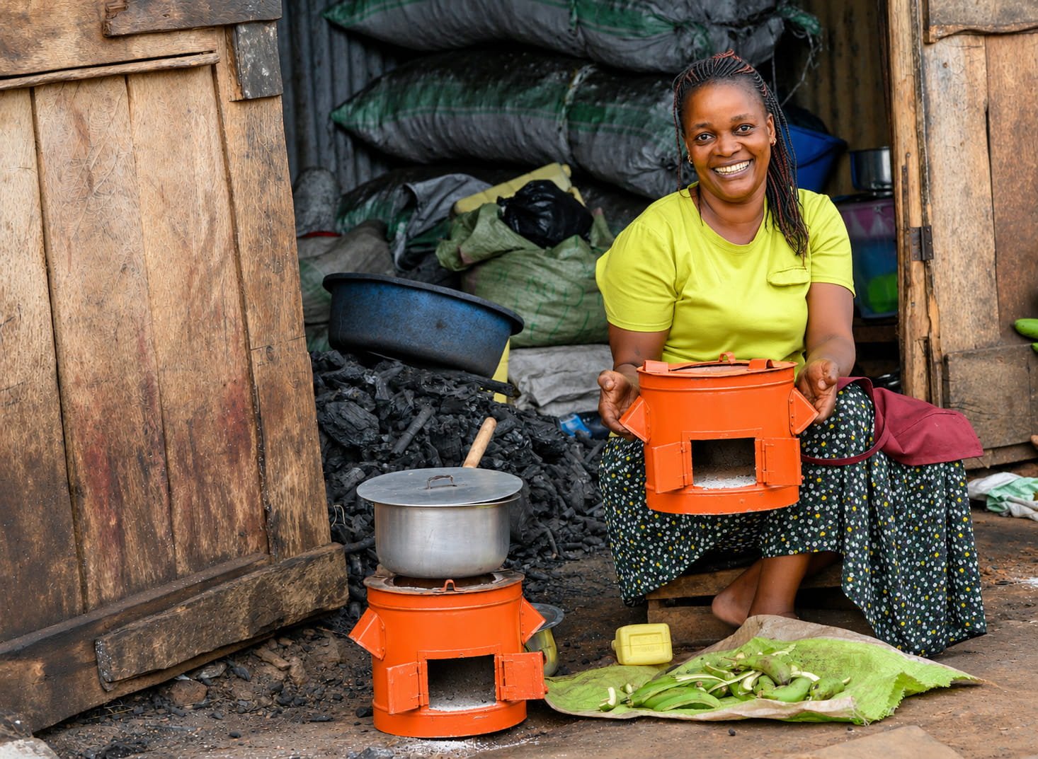 Smiling woman in a yellow shirt sitting on a small seat outside a wooden structure, holding an orange  impact carbon cookstove, with another stove nearby. 