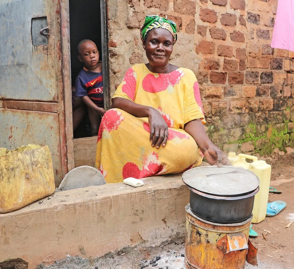 A woman in a yellow dress with red and pink pattern cooking over a impact carbon cookstove. A boy is standing in a doorway behind her, looking out. There are containers of fuel and cooking supplies around.