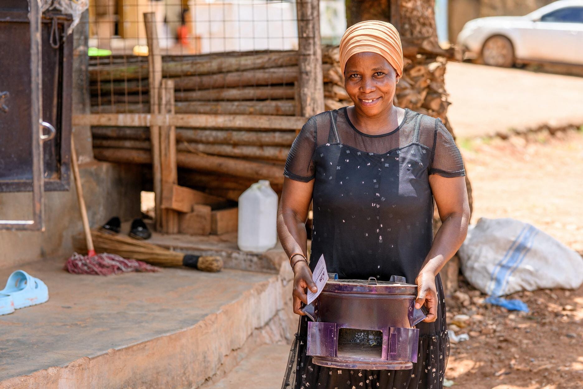 A woman standing outside holding a purple container with a stick attached to it. She is smiling and wearing a black dress with a peach-colored headscarf. In the background, there are tools, a white container, and a car parked on the dirt road.