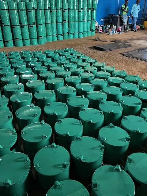 Many green water tanks arranged on the ground with a stack of similar tanks in the background, and two people standing near a table with supplies.
