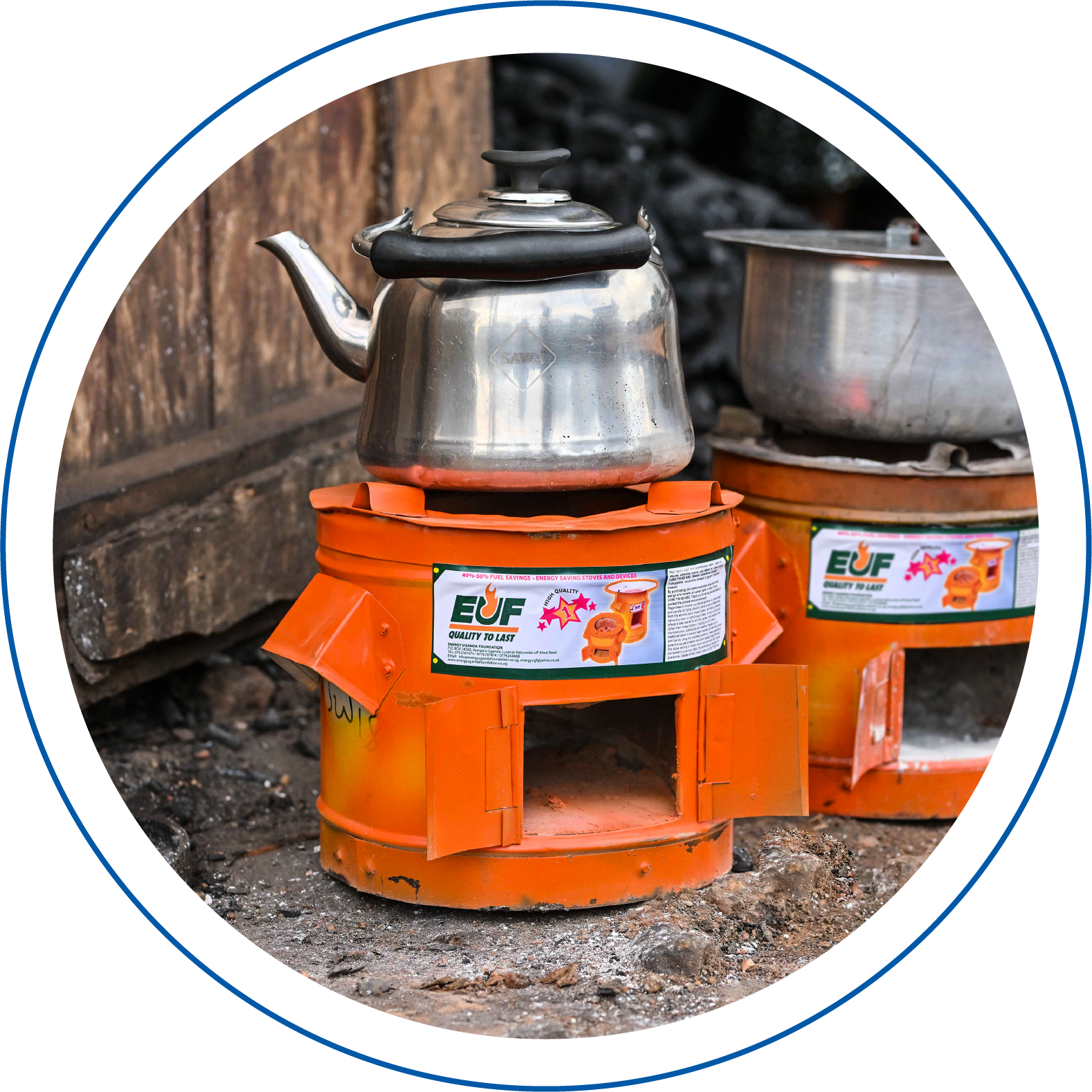 A steel kettle on a orange impact carbon stove with a label, and a large cooking pot on another stove in the background, set outdoors on dirt ground.