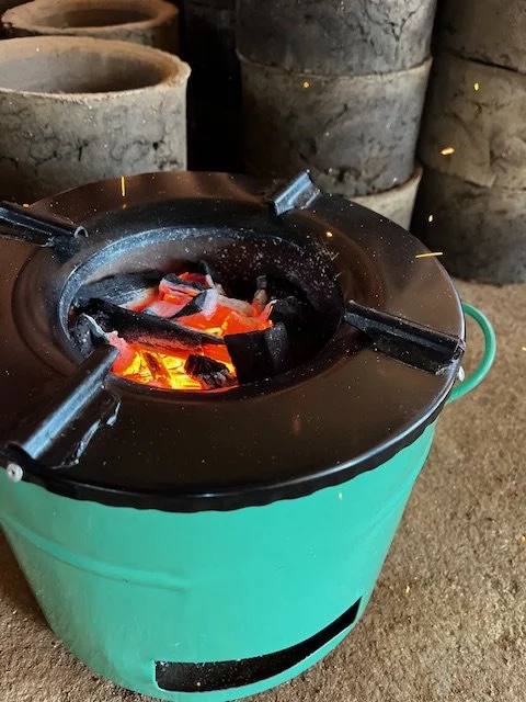 A small charcoal stove with glowing charcoal inside, used for cooking, with additional charcoal in the background.