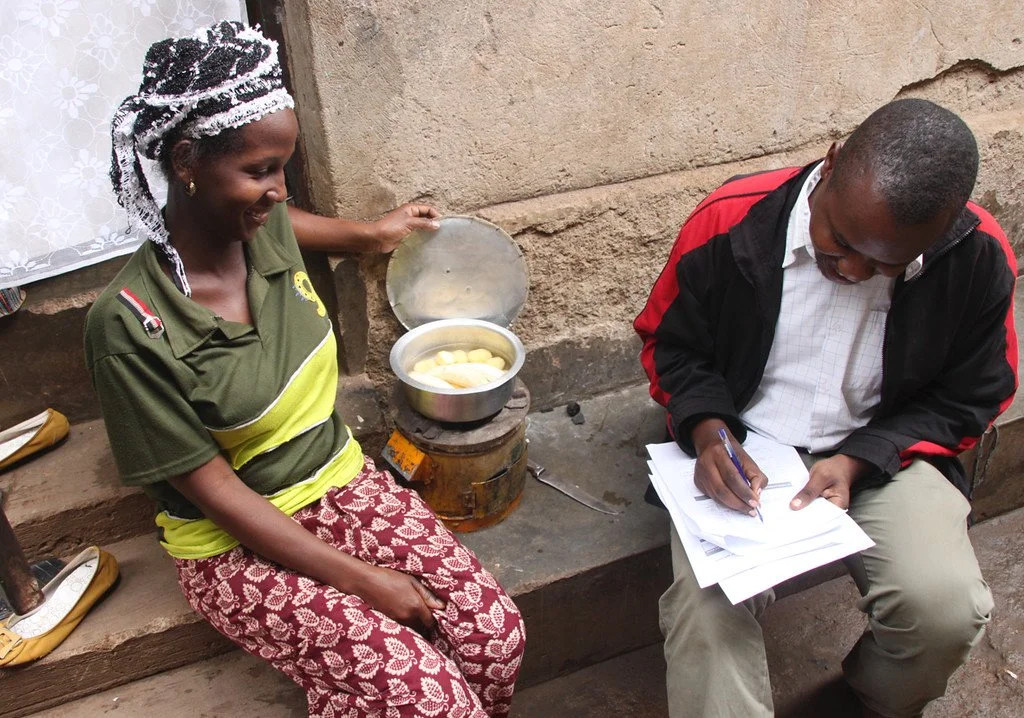 A woman sitting on a stone bench next to a man who is writing on a stack of papers. The woman is smiling and lifting a metal container with a lid, containing boiled potatoes. They are outdoors against a rough wall.