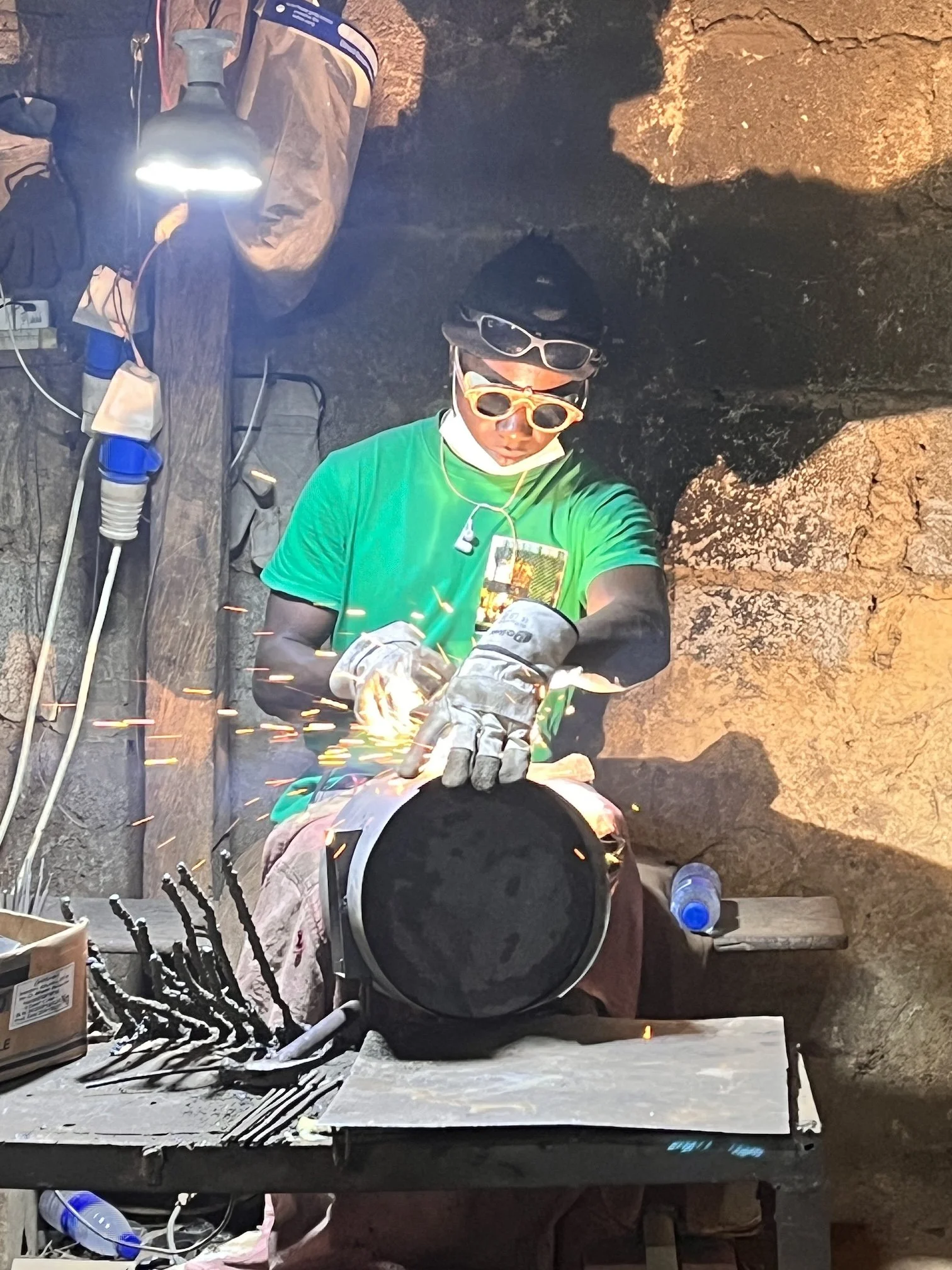 Welding work happening inside a rocky cave, with sparks flying from the welding process. A worker wearing protective gear, gloves, and sunglasses is focused on welding a piece of metal. Tools, supplies, and equipment are scattered on a table nearby.