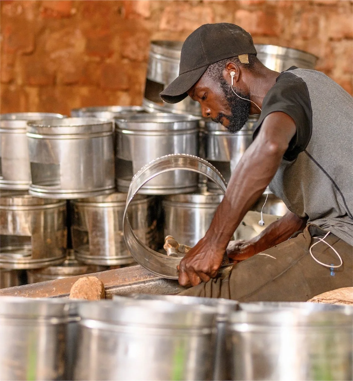 A man working on a metal drum with a hammer, surrounded by stacked improved cookstoves in an industrial setting.