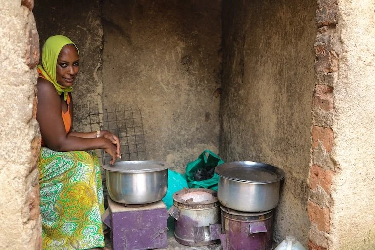 A woman in a colorful dress and green headscarf sitting in a small, rustic kitchen with pots and cooking appliances.