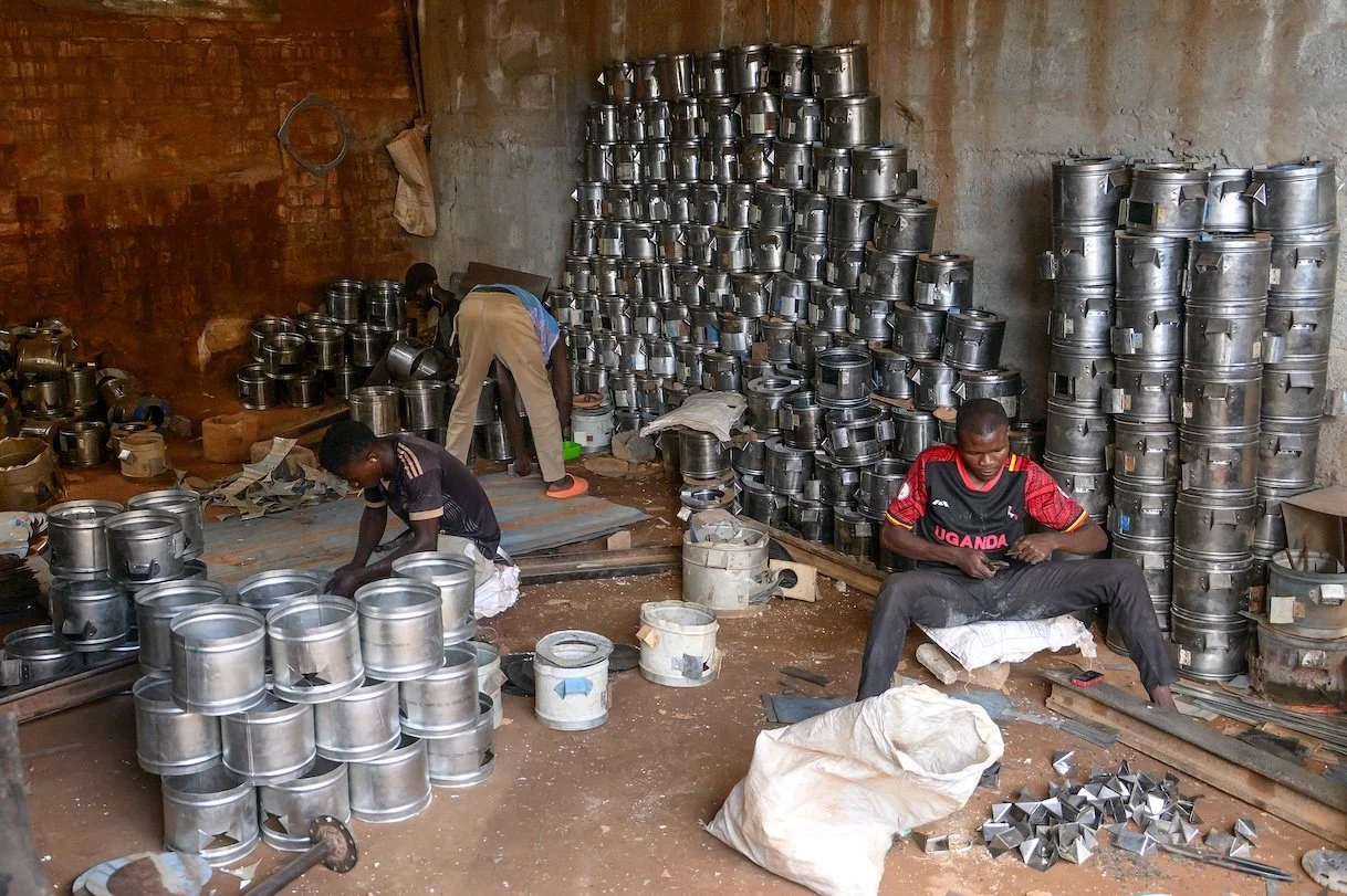 Three men working in a metal workshop surrounded by stacked metal pots and lids, with a brick and concrete wall in the background.