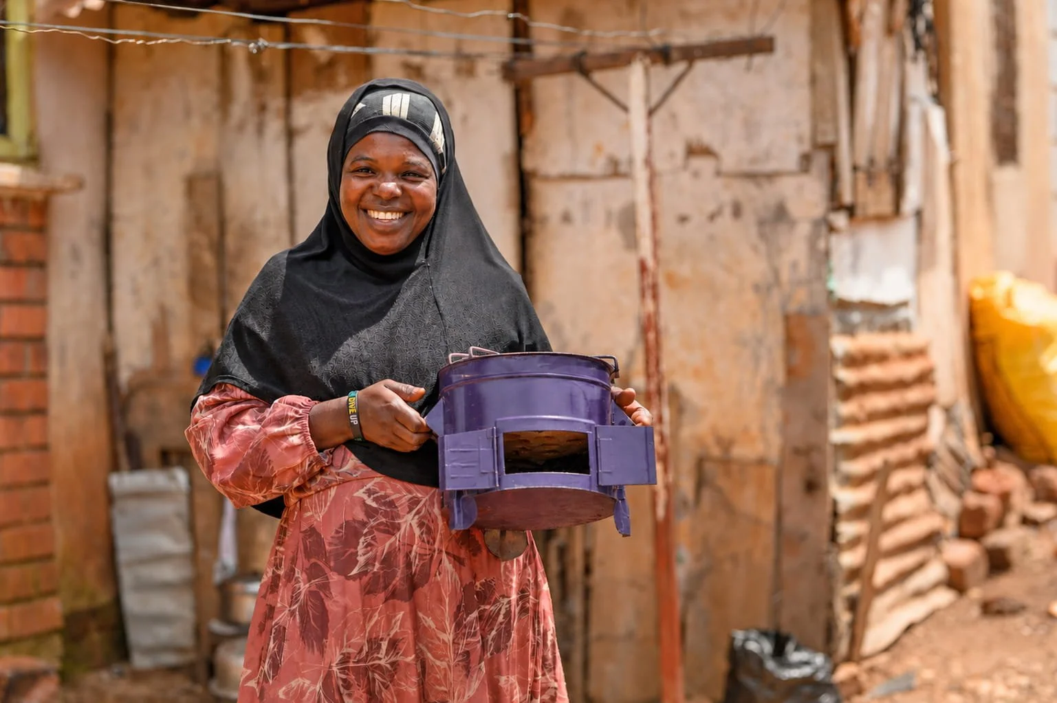 A woman smiling and holding a purple stove in a rustic, unfinished building with brick and wood walls.