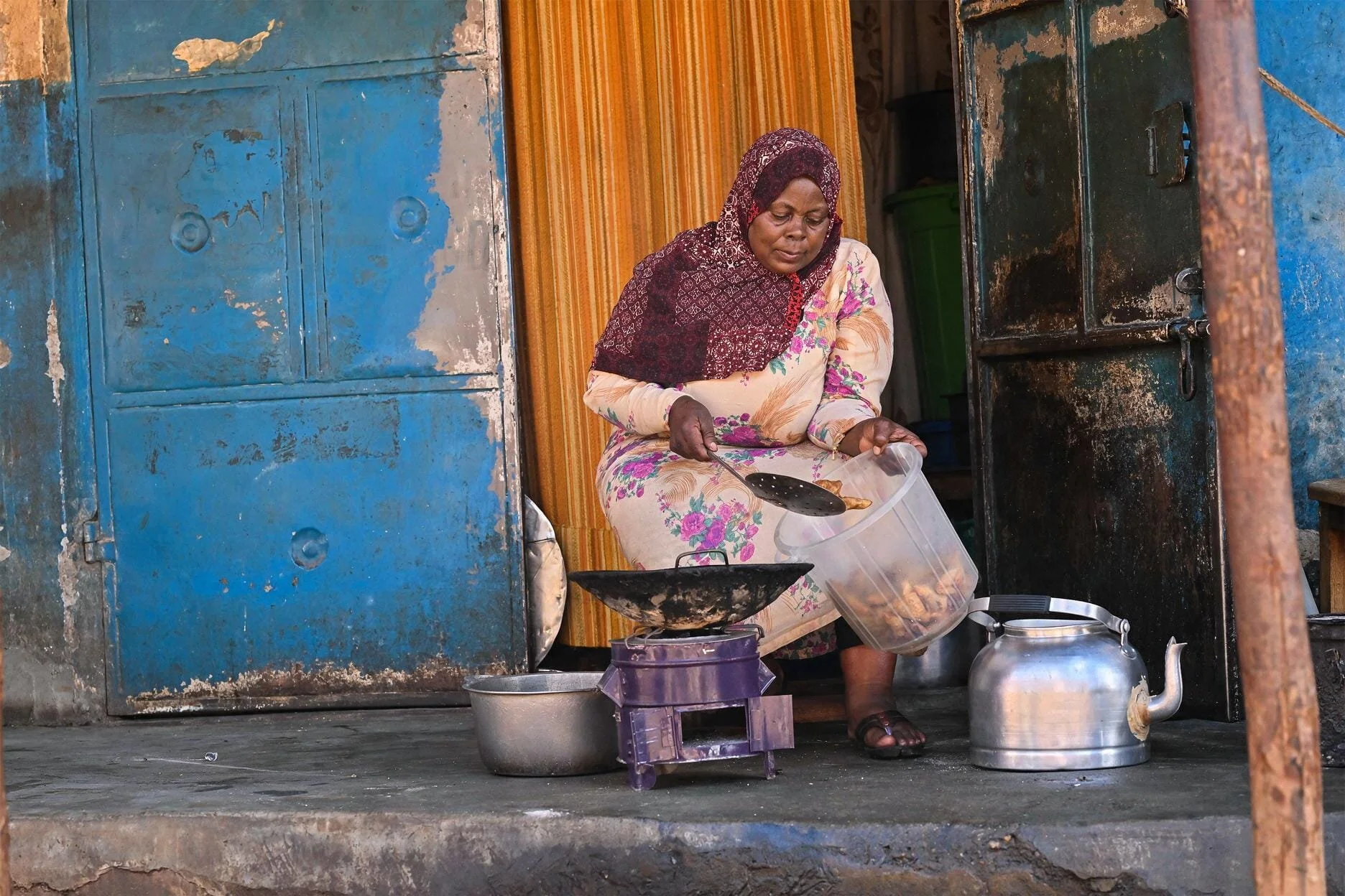 A woman in traditional clothing cooking outside a small shop or home, sitting on a low stool, pouring food from a large plastic container into a pan on a small stove.