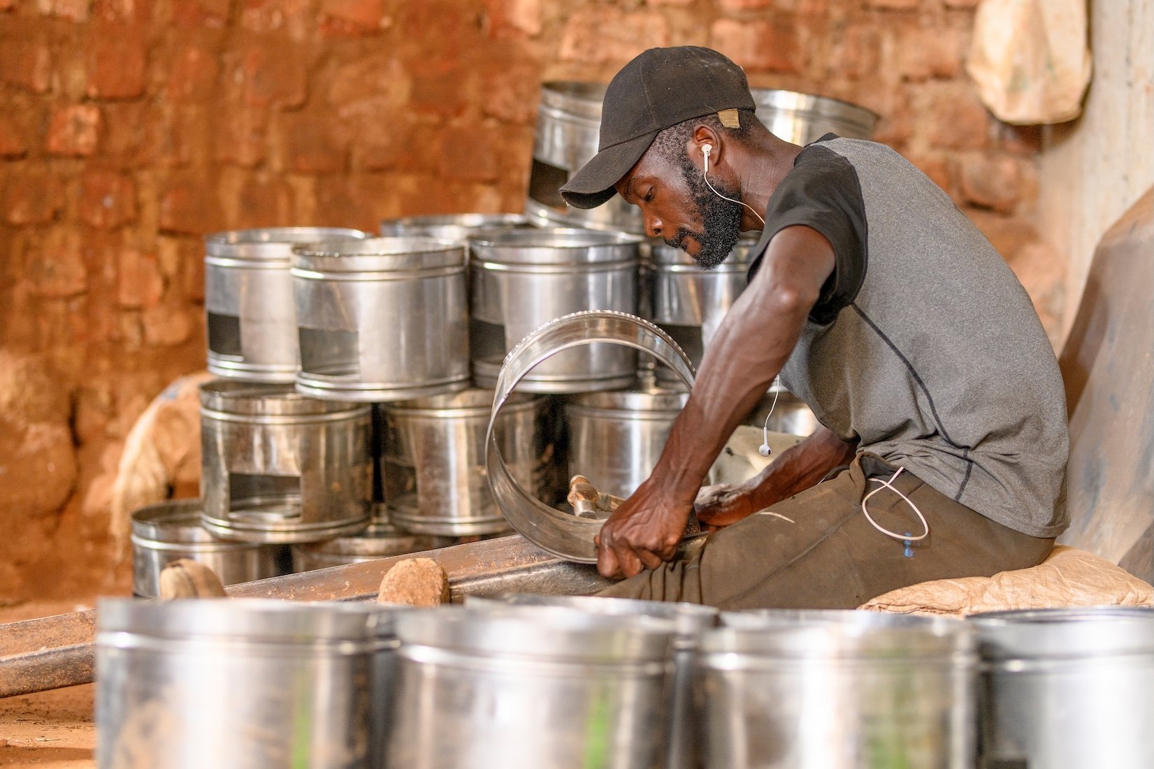 A man wearing a black cap, gray T-shirt, and brown pants is sitting on a cushion, working on a large metal ring with a hammer inside a workshop with a brick wall background. Several metal cans are stacked behind him, and there are some tools and mate