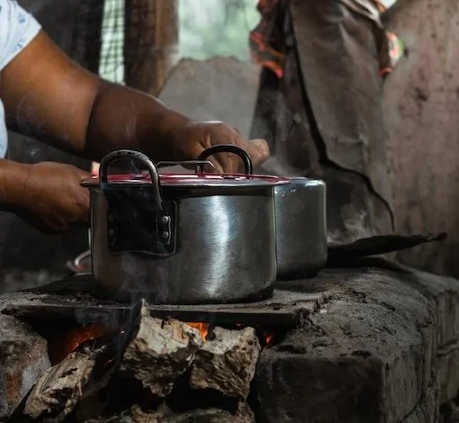 Two traditional cookstovs on  an open flame, with a person's hands handling one of the pots.