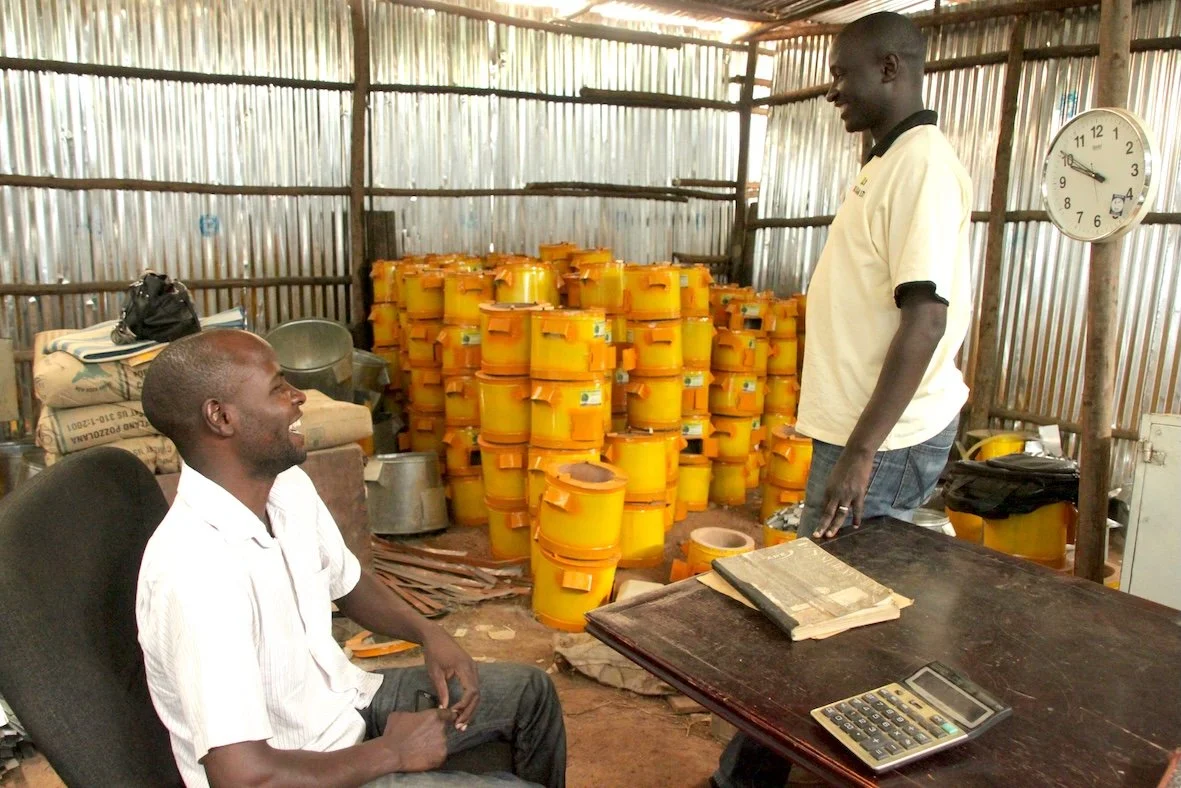 Two men talking inside a small workshop with impact carbon cookstoves stacked in the background.